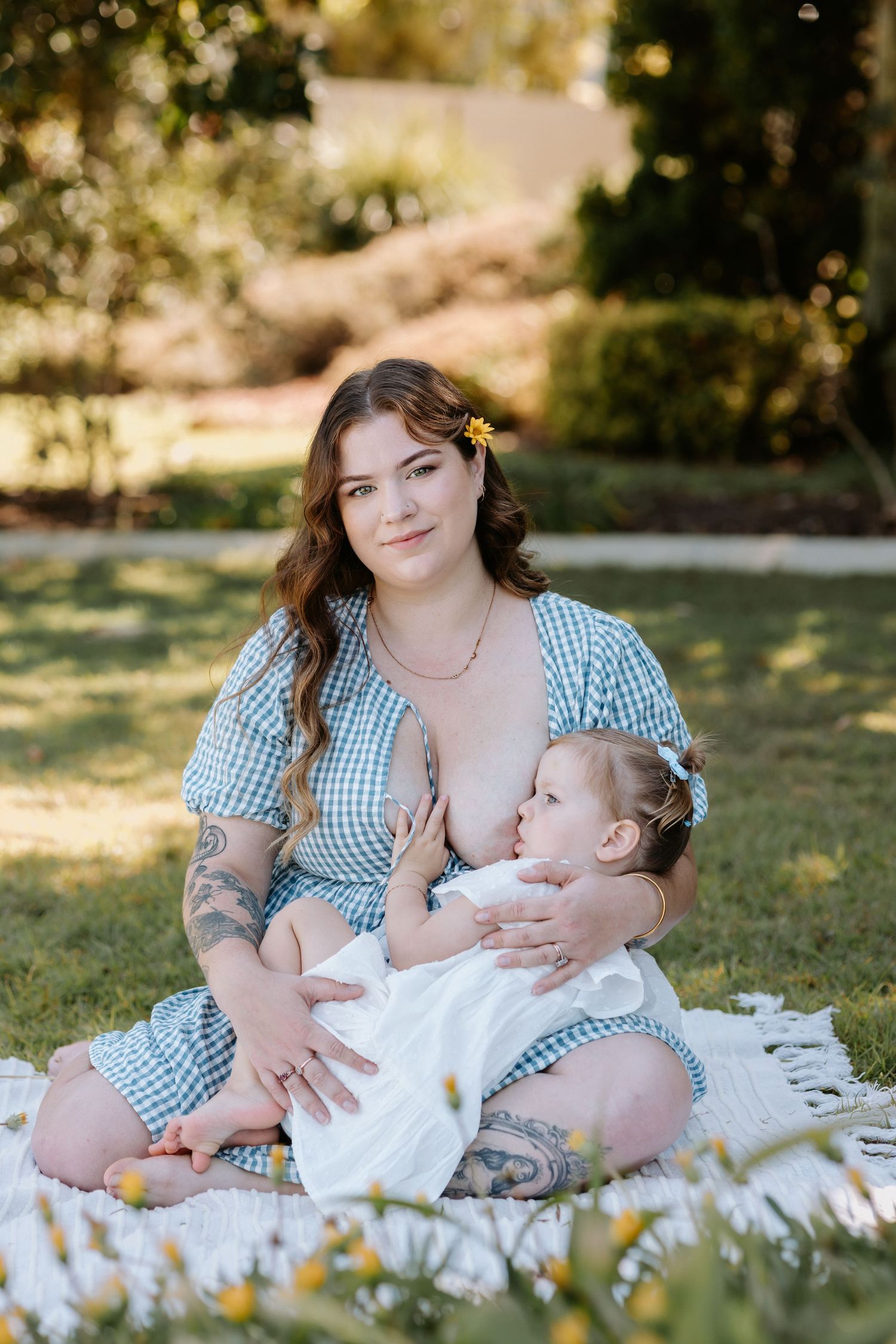 Mother and baby share tender breastfeeding moment on blanket in sunny garden wearing matching gingham outfits.