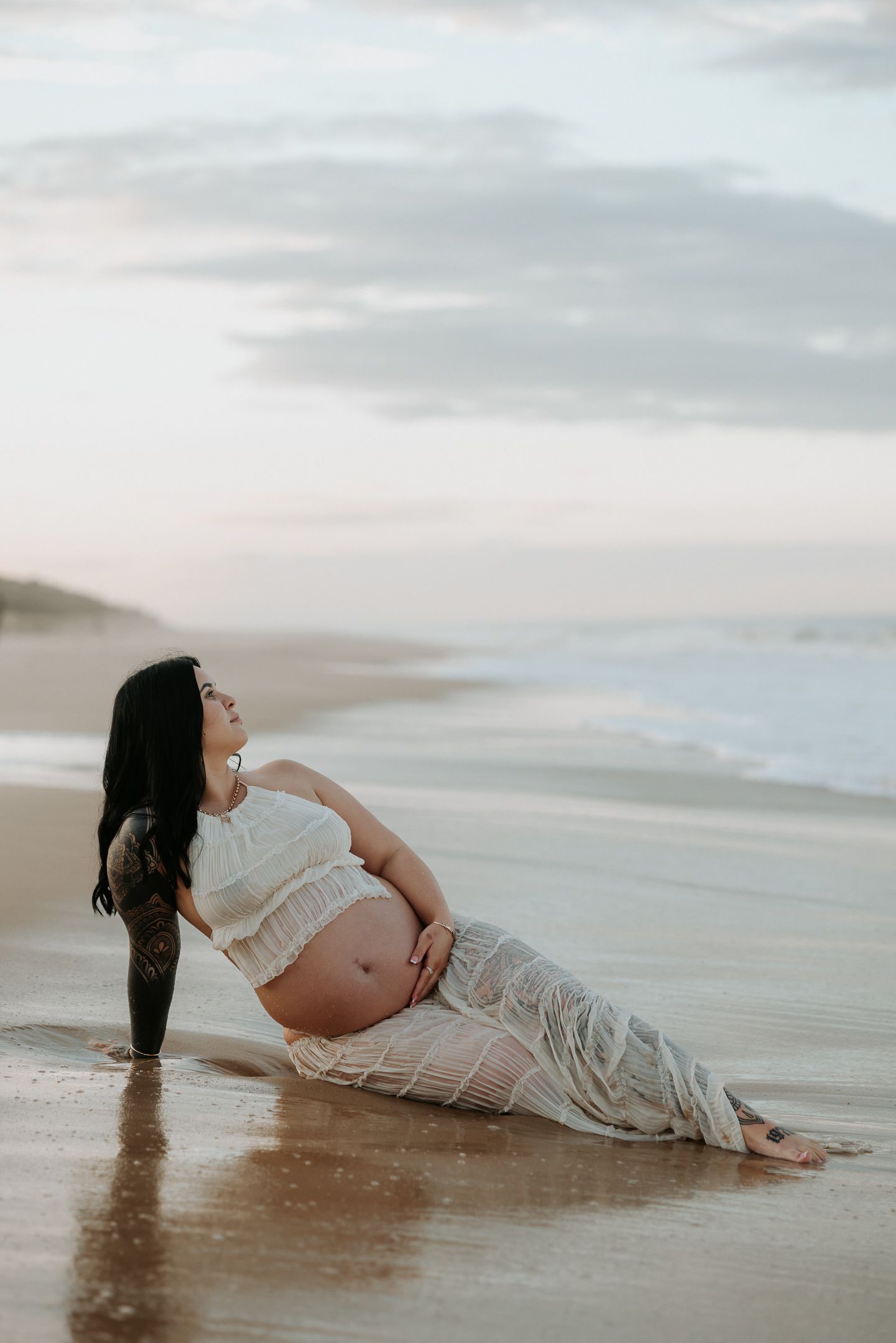 Artistic maternity photoshoot showing a woman in white dress lying in shallow ocean water at sunset.
