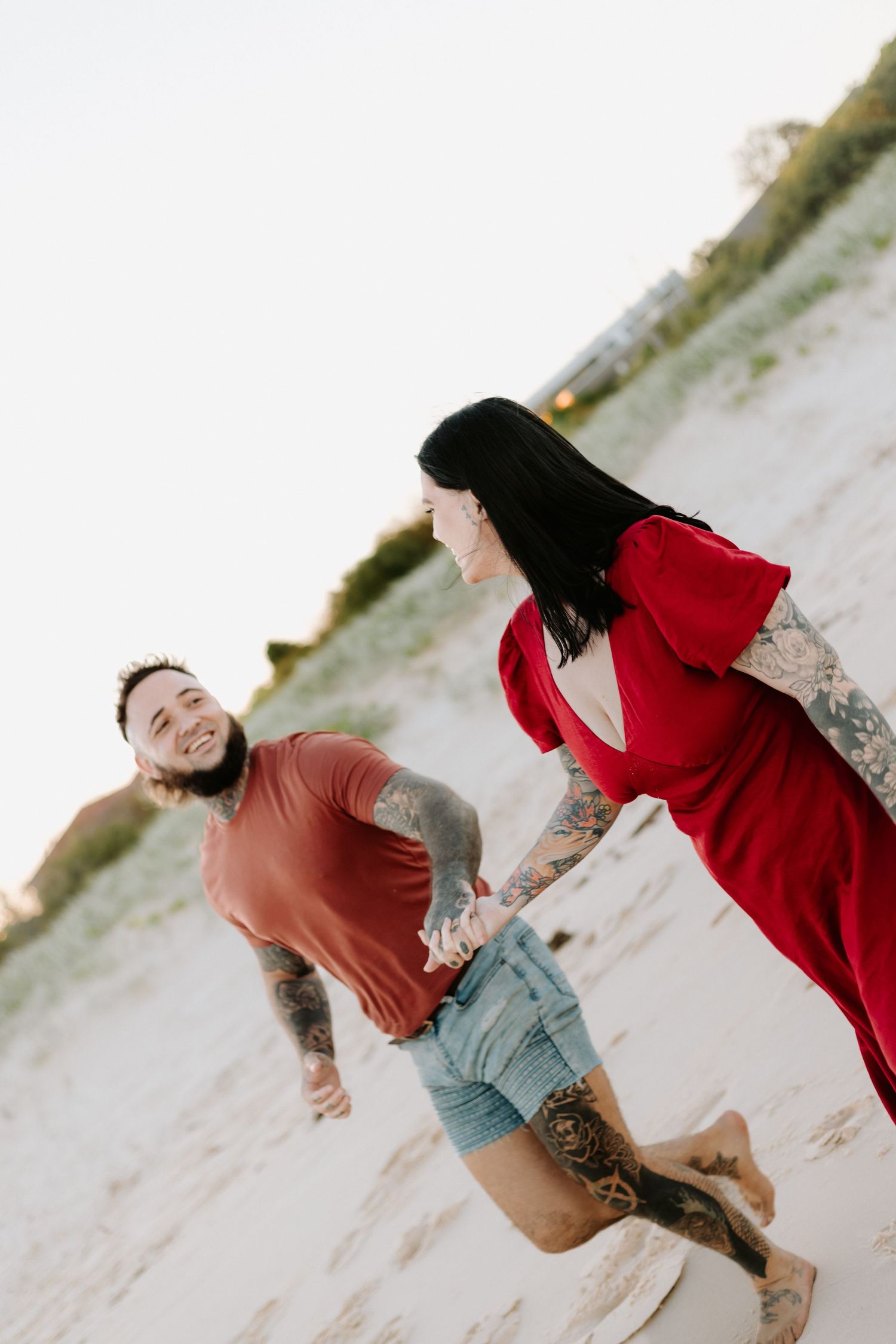 A couple in red and brown outfits playfully dance and embrace on a sandy beach at sunset.