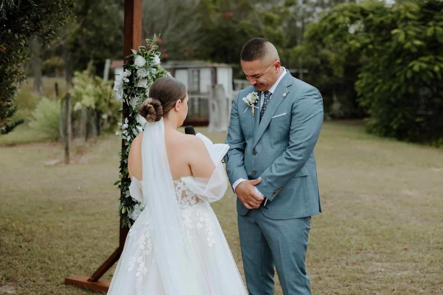 Bride and groom share an intimate first look moment in a lush garden setting wearing blue suit and white gown.