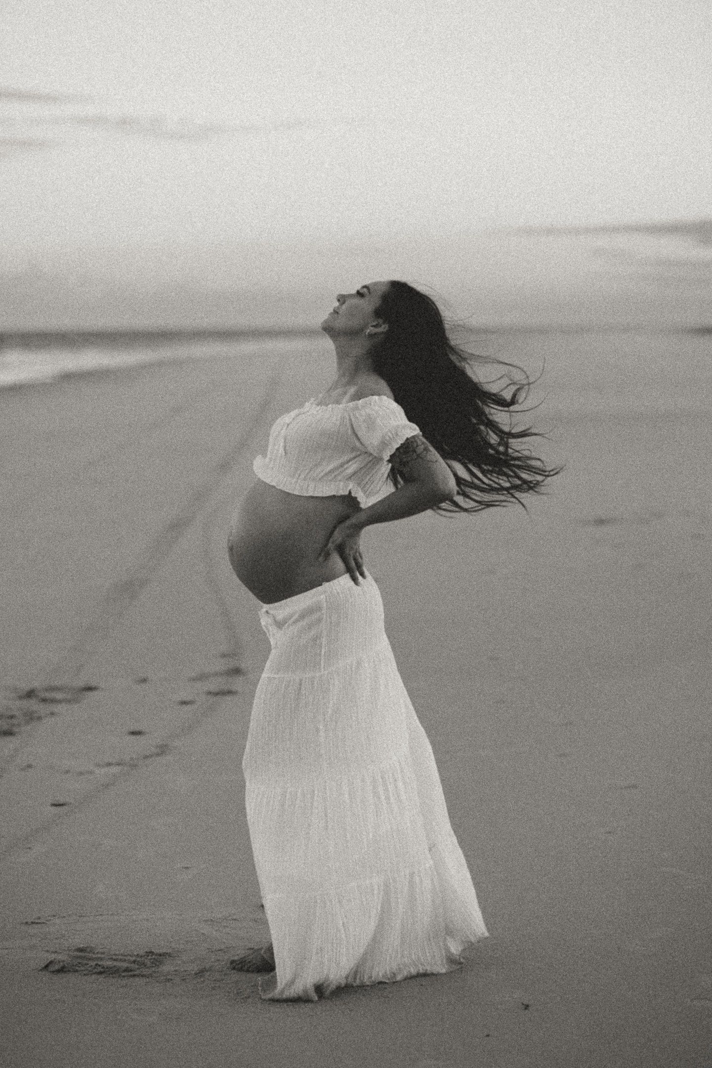 A figure in a flowing white dress dances gracefully on a misty beach in black and white photography.