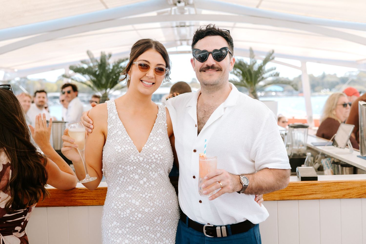 Couple smiling at an outdoor beach bar event, woman in white dress and man in white shirt with sunglasses.