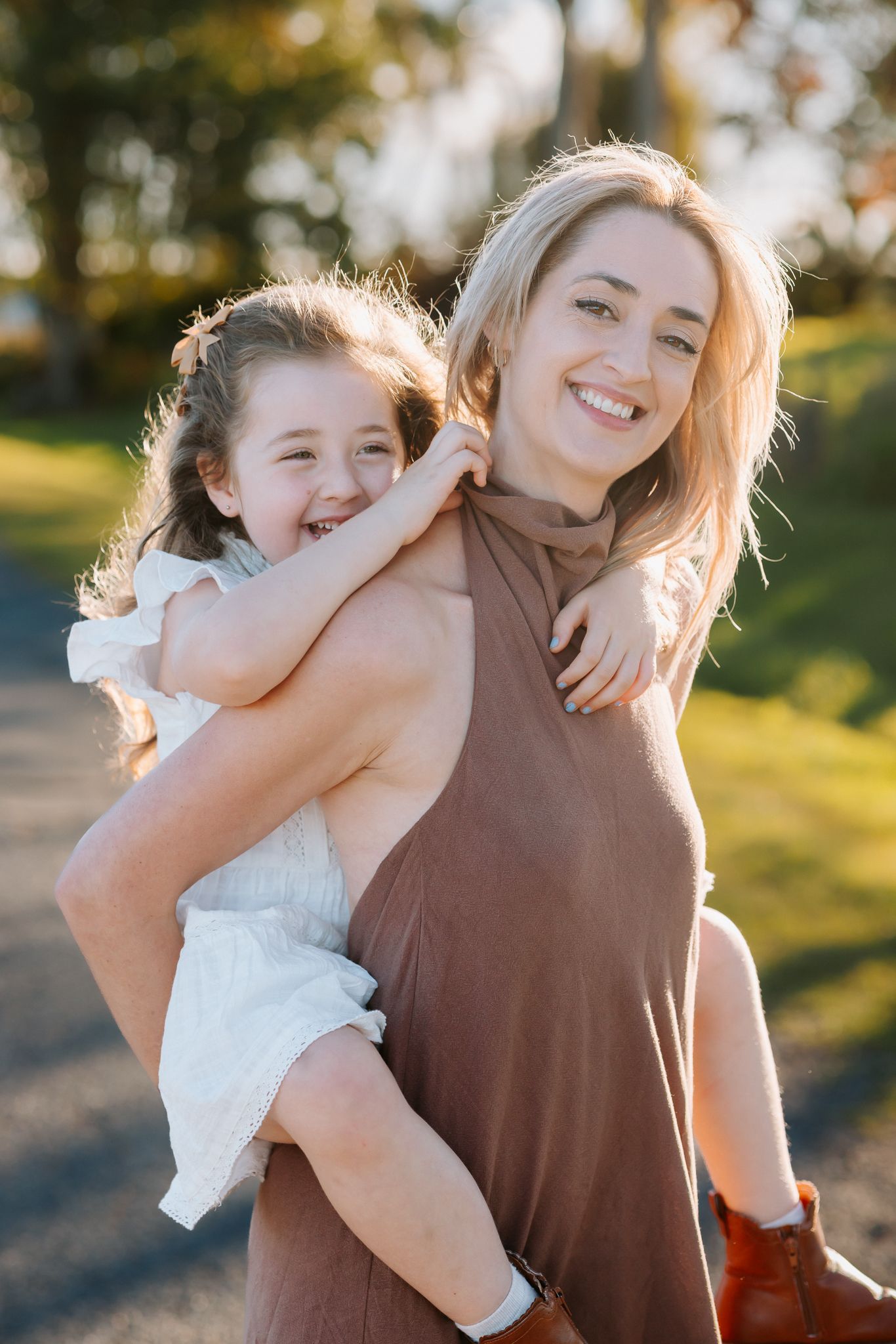 A smiling woman in a brown dress carries a laughing little girl on her back outdoors in warm sunlight.