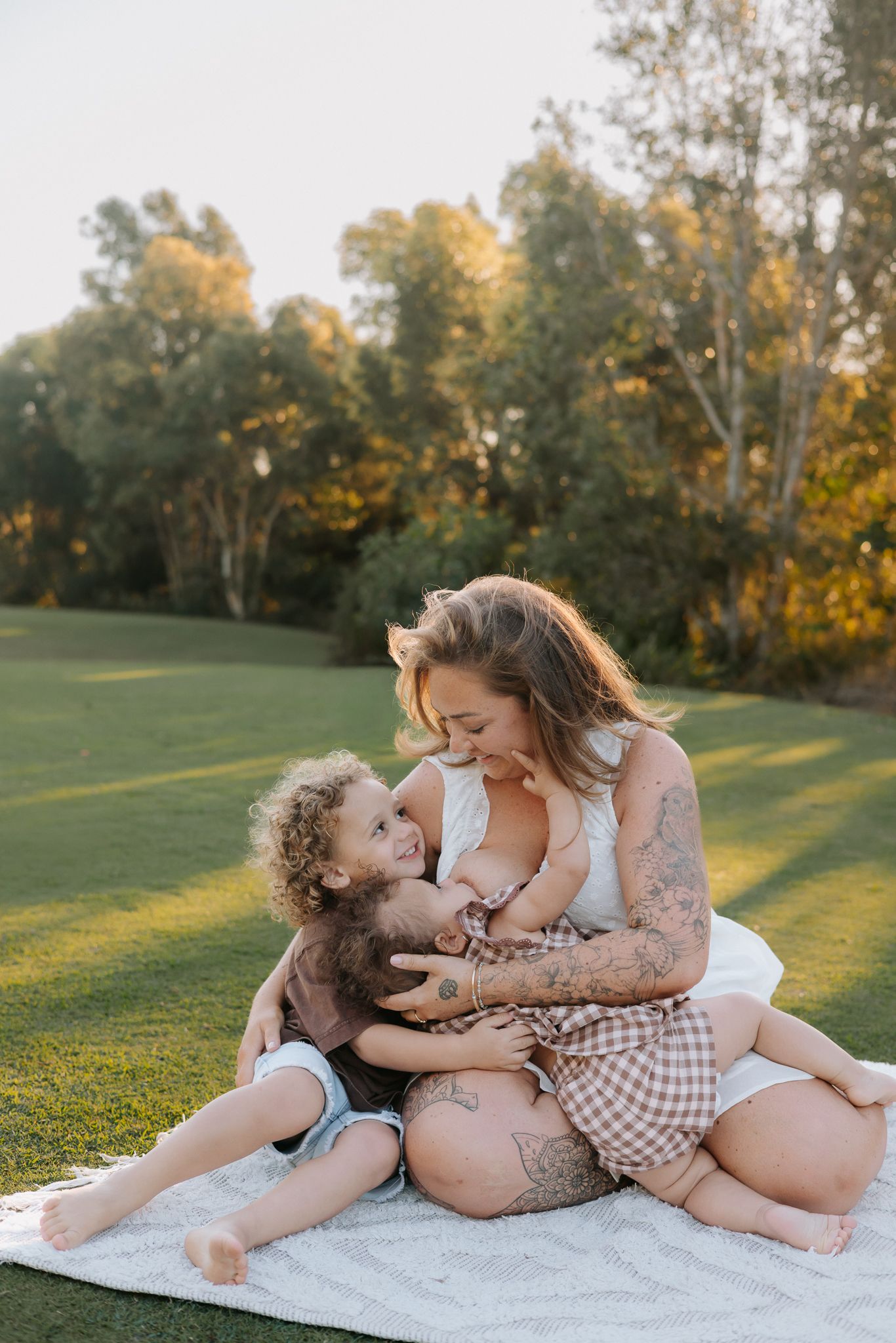 A tattooed mother sits on a blanket in a sunny park, lovingly embracing her two young children outdoors.