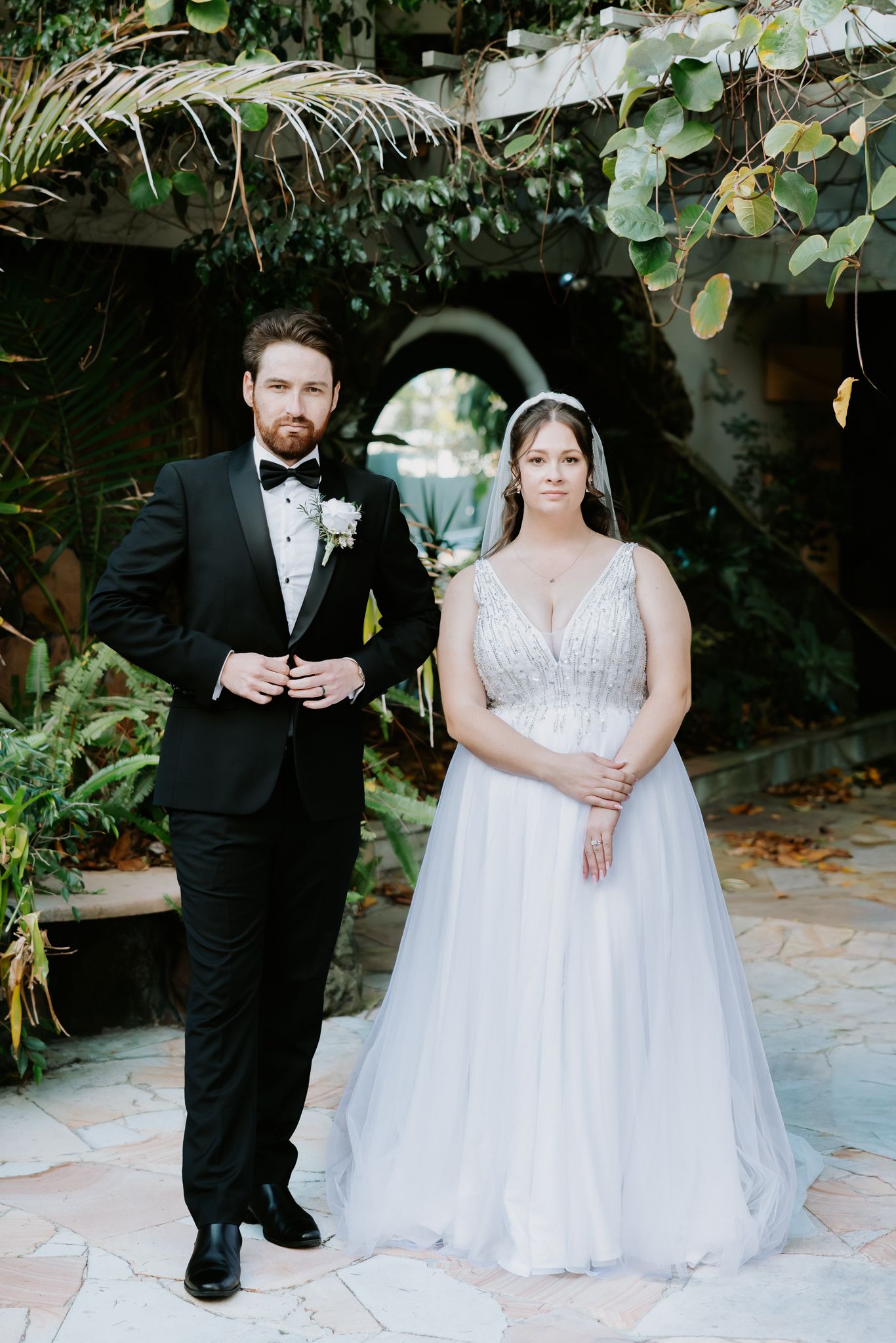 Bride in beaded ballgown and groom in black tuxedo pose together in lush garden courtyard on their wedding day.
