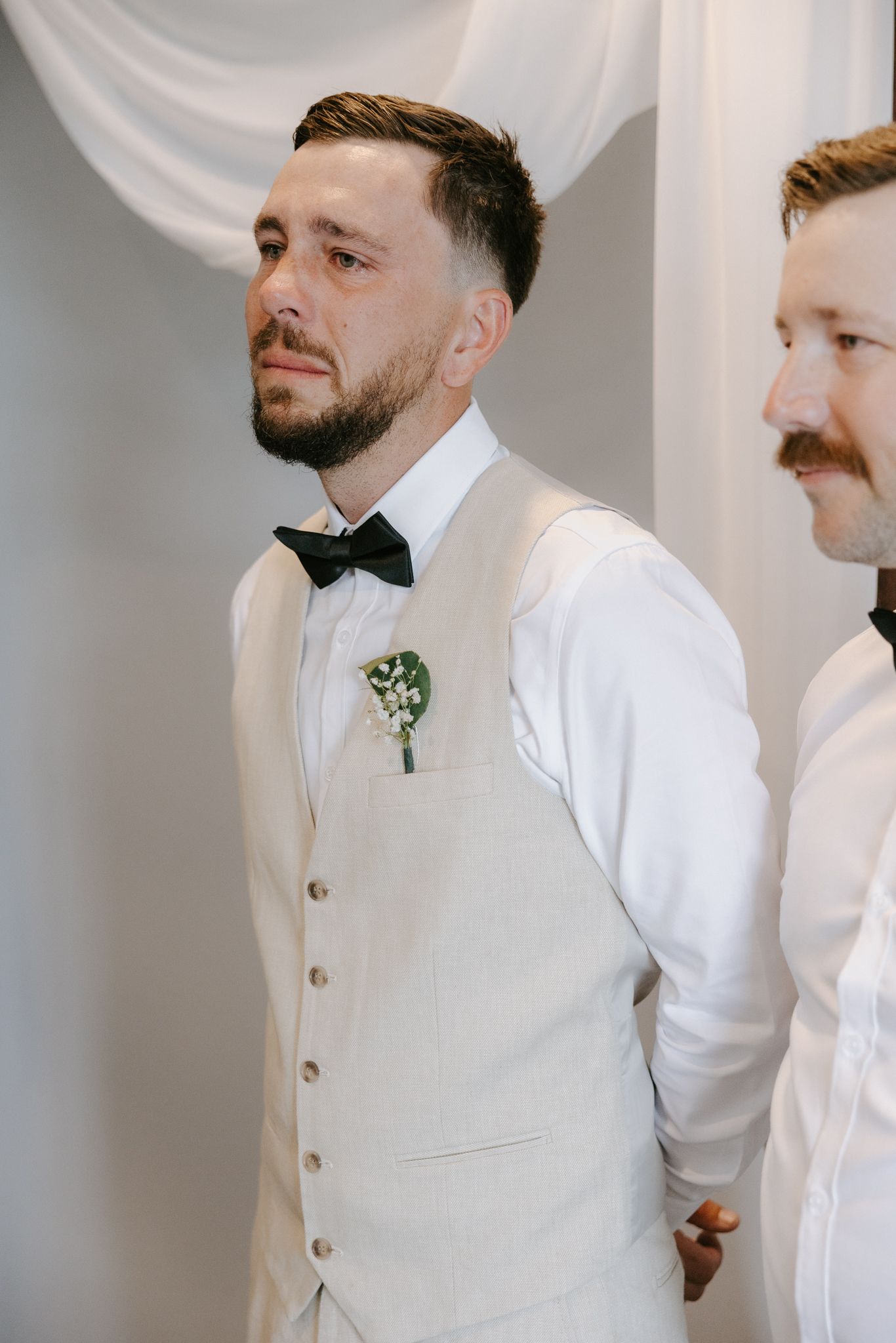 Emotional groom in beige vest and bow tie crying during wedding ceremony with boutonniere on lapel.