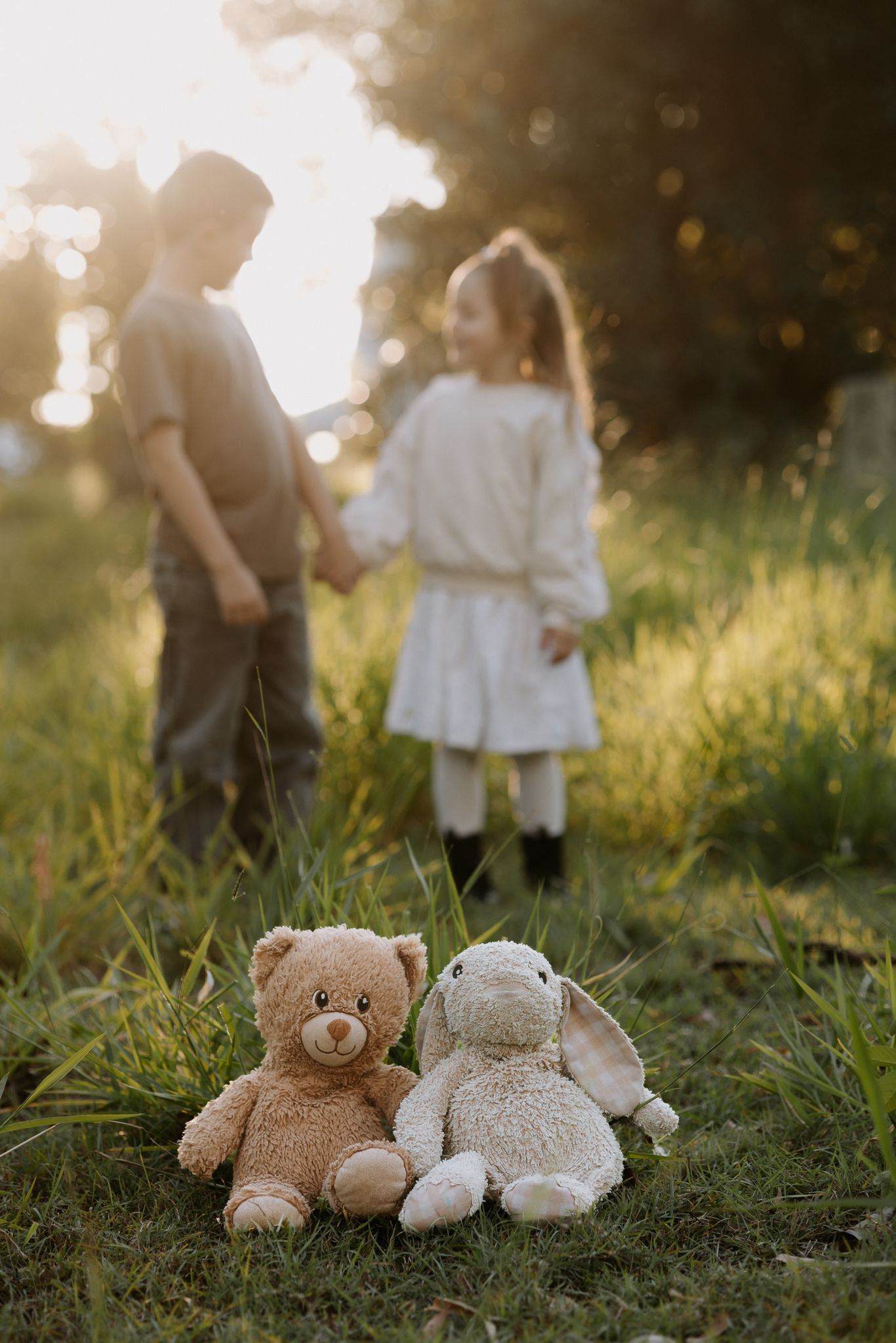 Two children hold hands in a sunlit meadow with teddy bear and bunny plush toys sitting in the foreground grass.