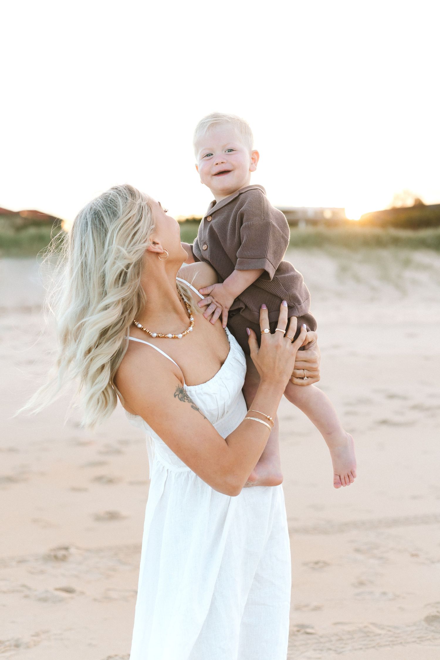 A blonde woman in a white dress holds a laughing baby boy on a sandy beach at golden sunset.