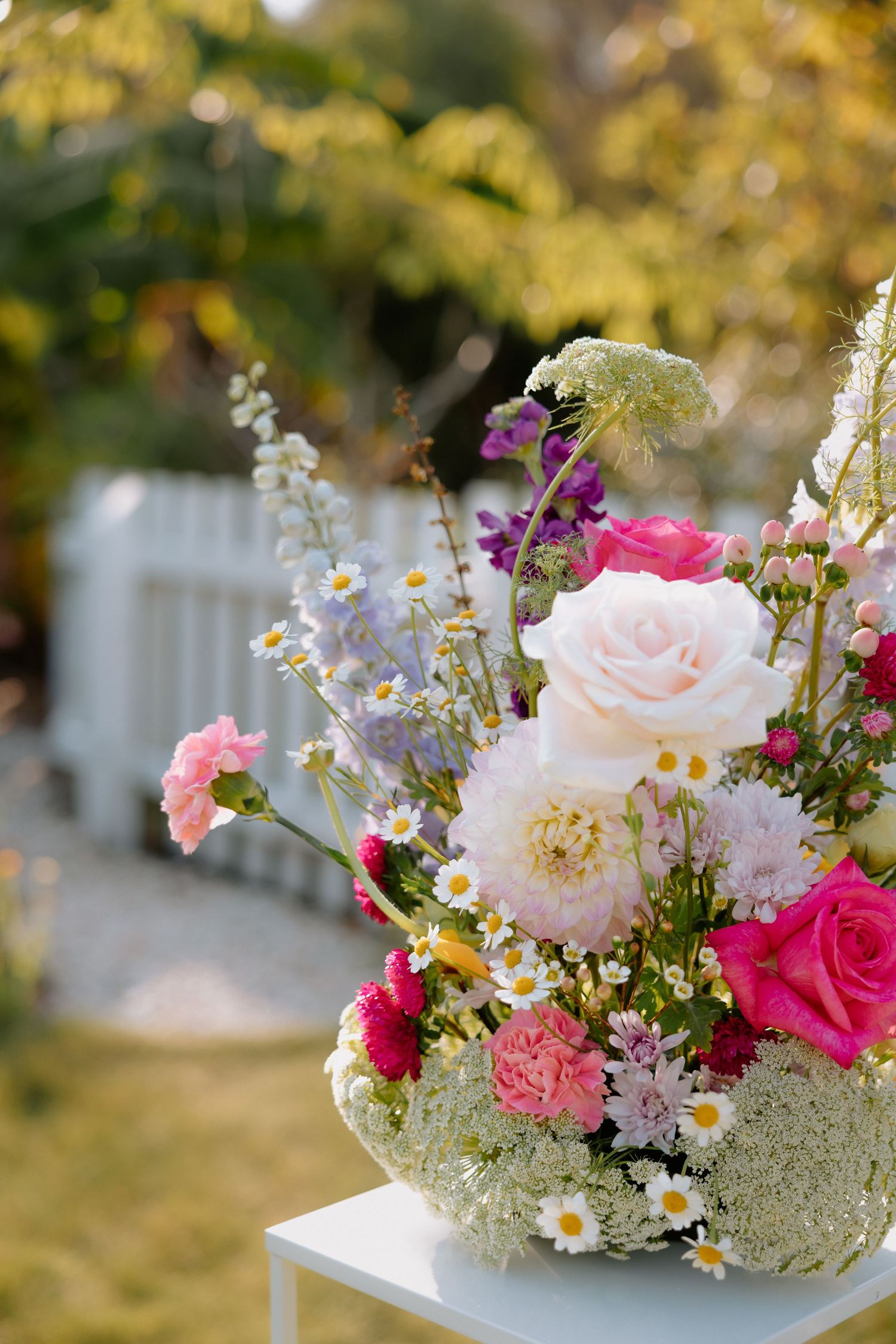 Lush outdoor floral arrangement featuring pink roses, daisies, and wildflowers in a garden setting with soft bokeh background.