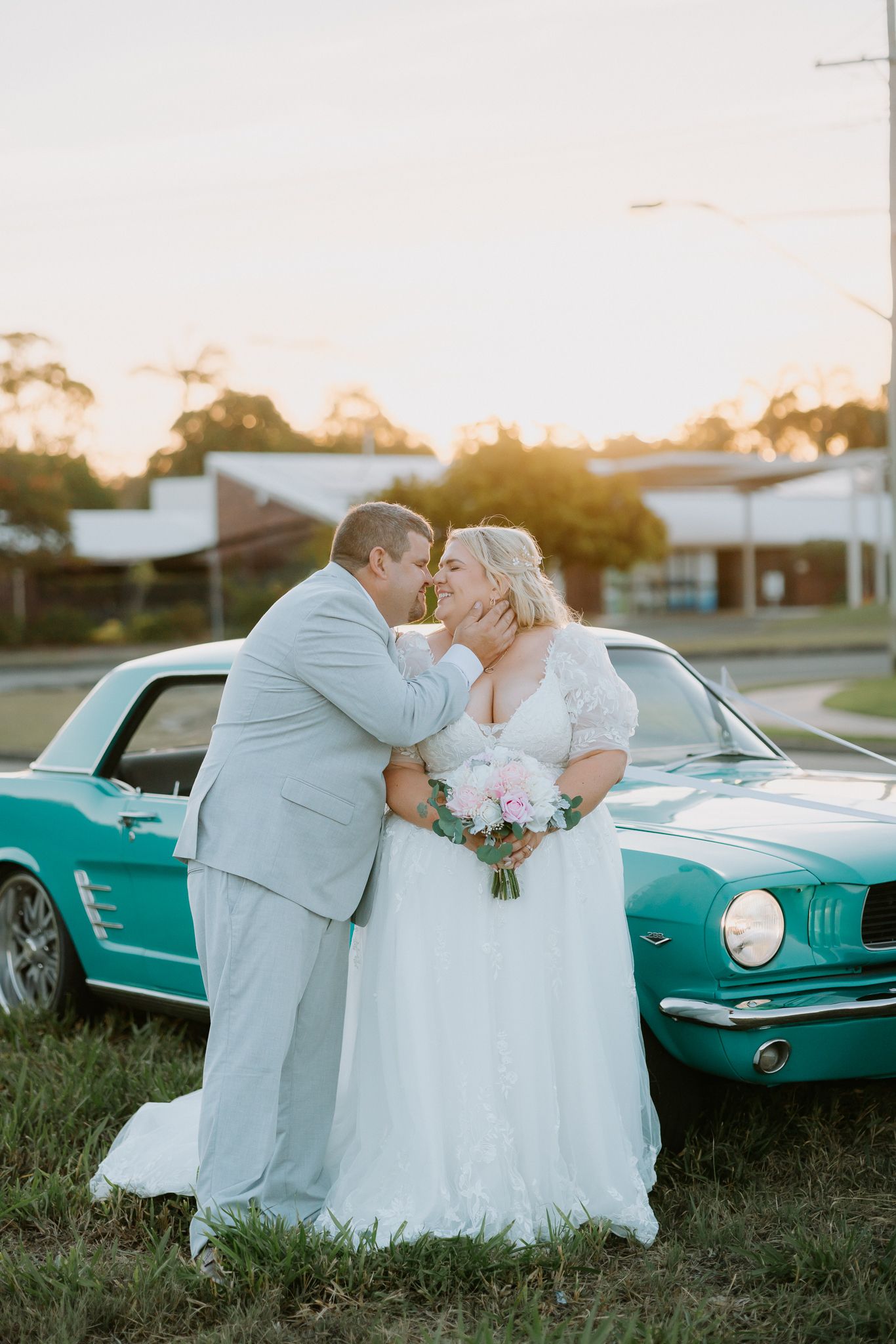 Bride and groom share a romantic moment in front of a teal vintage Ford Mustang at sunset.