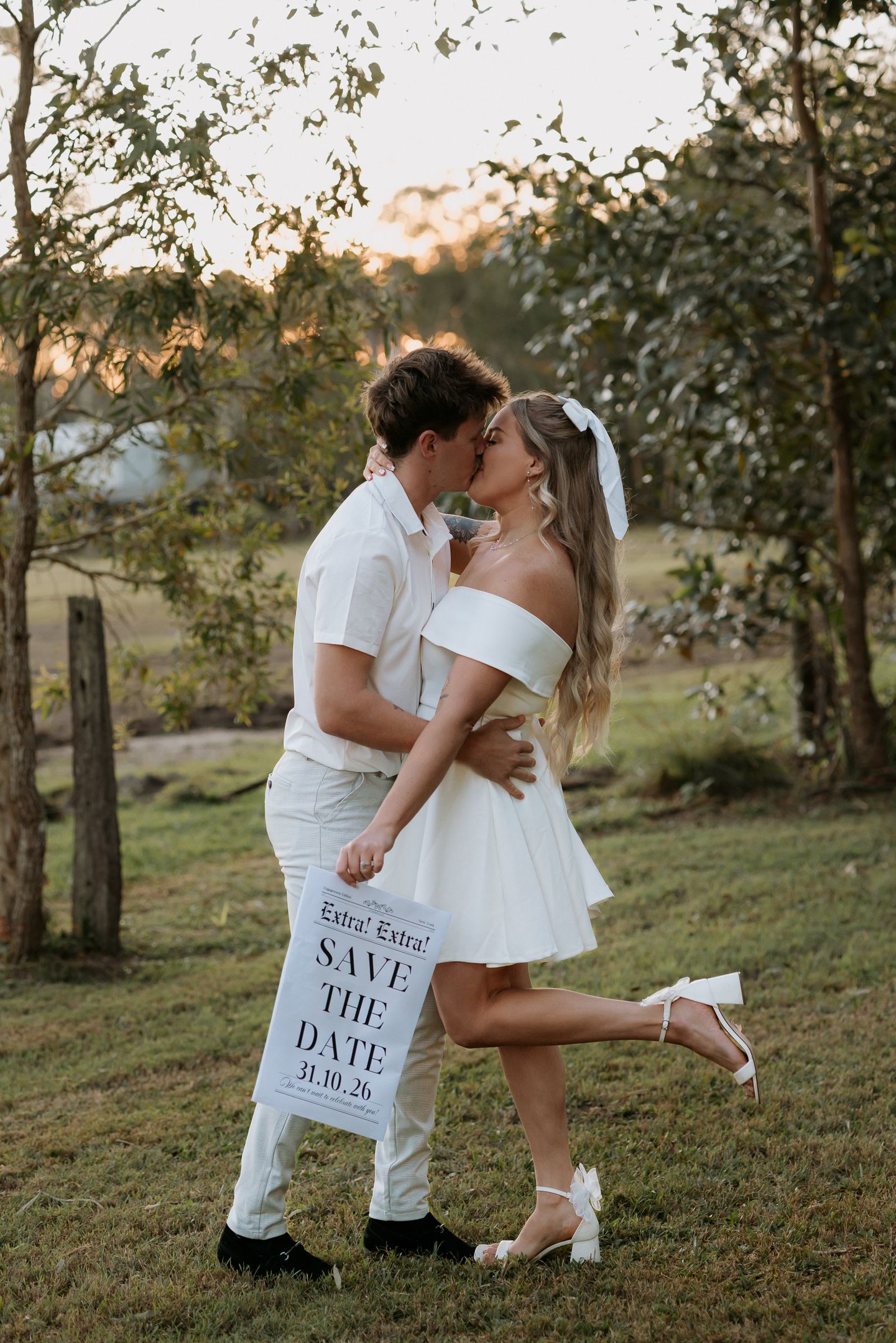 Couple kissing outdoors holding a Save the Date sign dated 31.10.26, bride in white mini dress and groom in white outfit.
