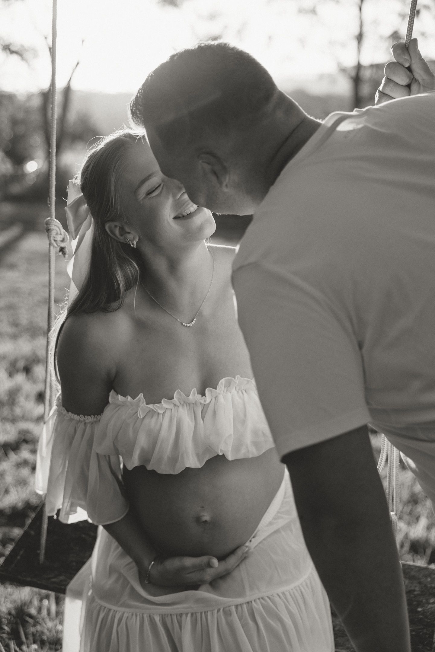 Black and white maternity photo of couple sharing tender moment, man kissing pregnant woman on forehead outdoors.