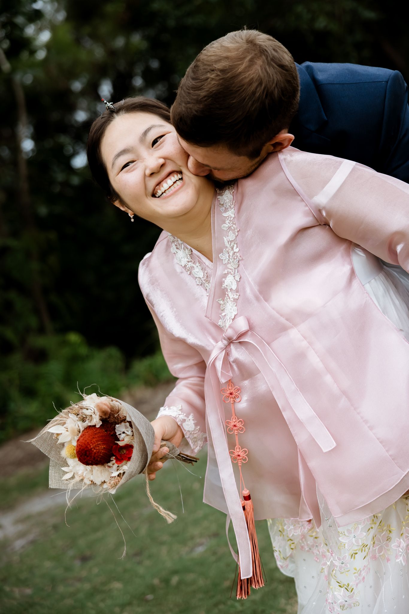 Joyful bride in pink hanbok laughs as groom kisses her cheek, holding a bouquet outdoors.