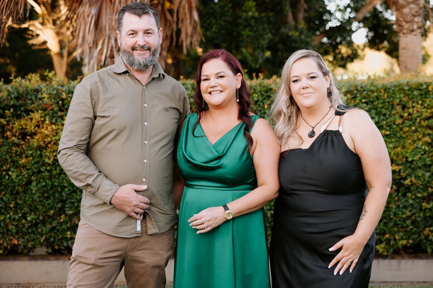 Three adults smiling outdoors, man in khaki, woman in green dress, woman in black dress.