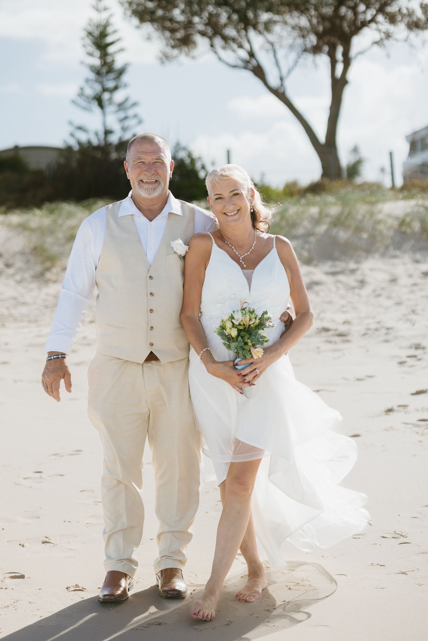 Happy couple in wedding attire walking barefoot on a sunny beach, bride holding bouquet in short white dress.