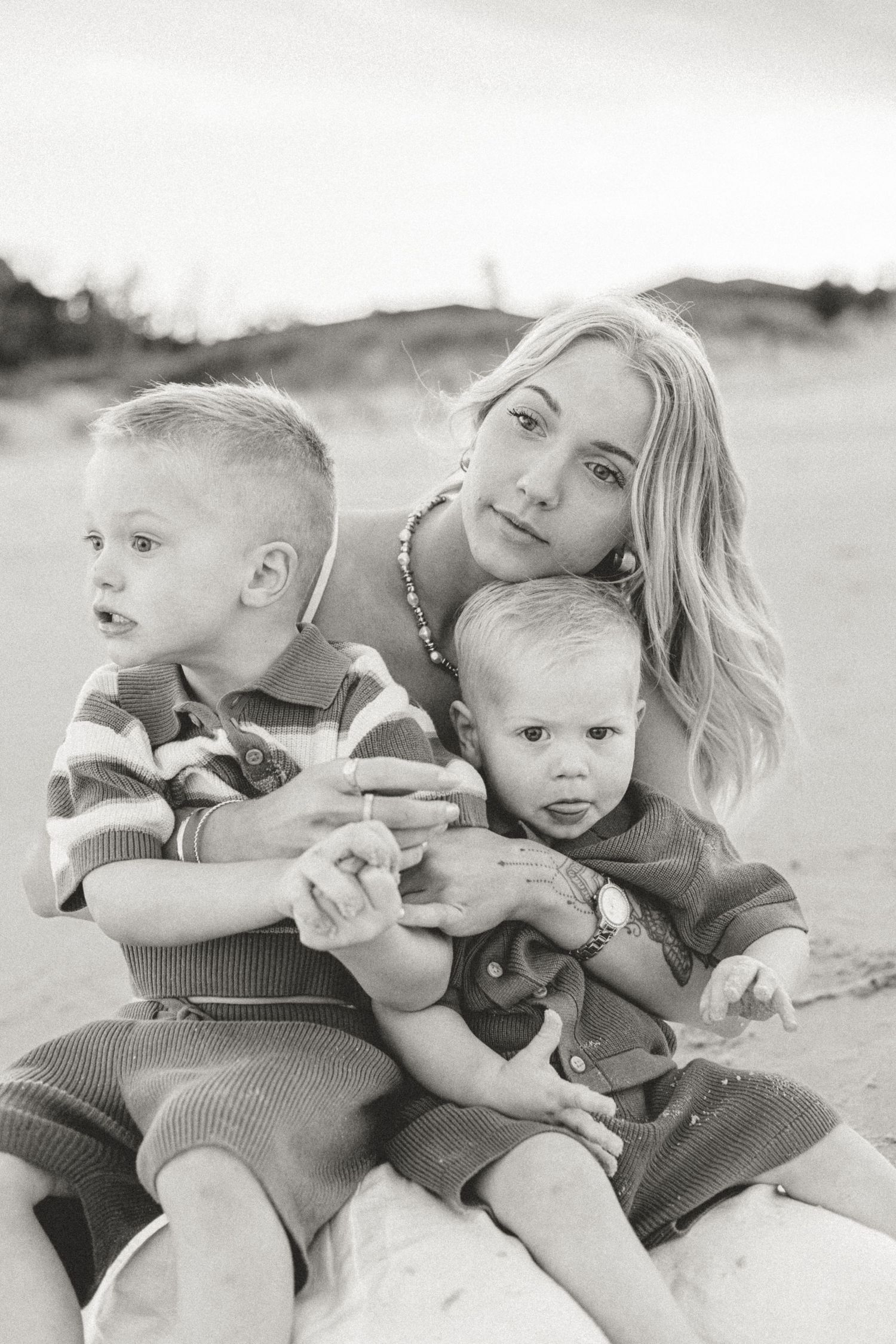 Black and white photo of a young mother sitting on a beach holding two small boys in her arms.