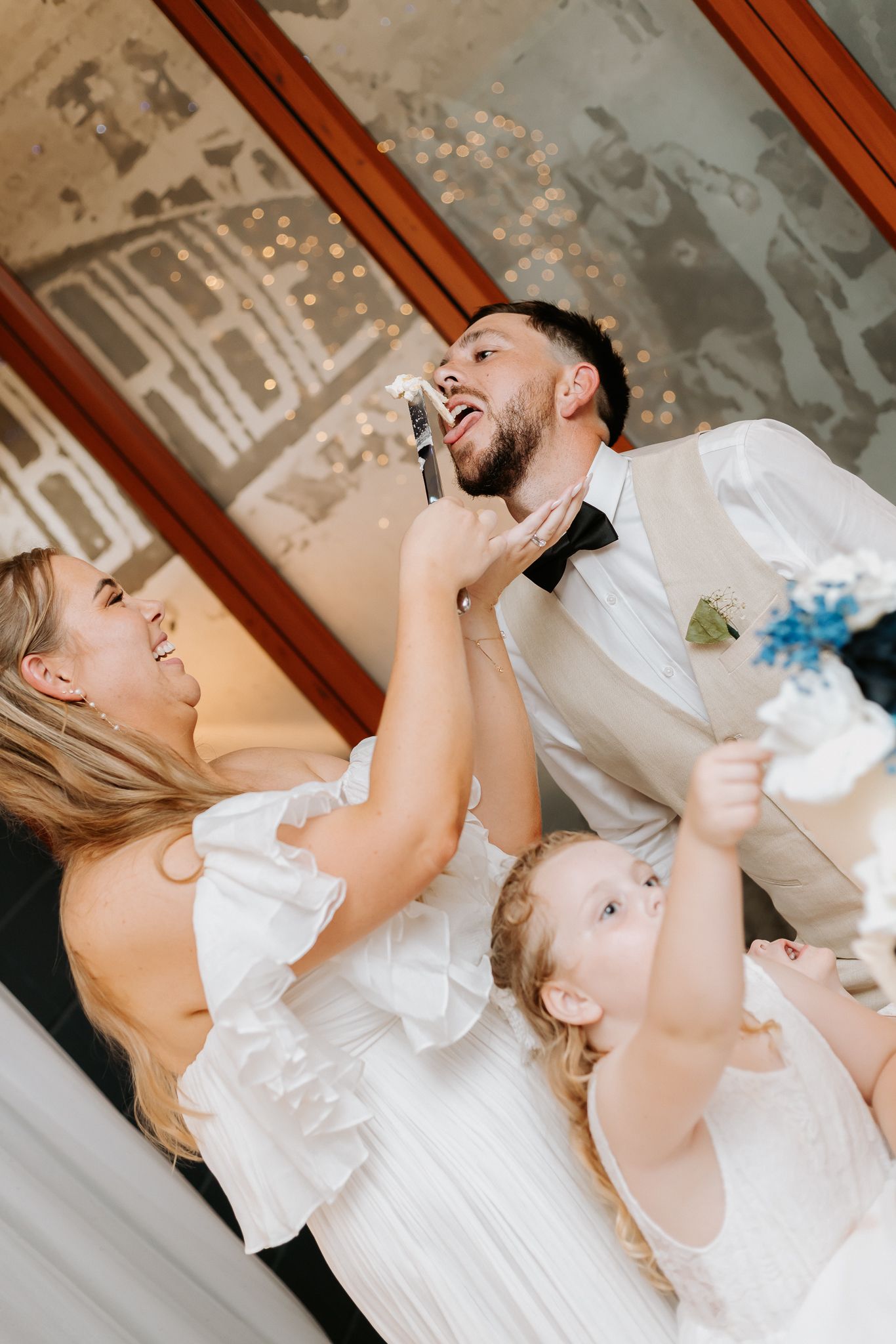 Bride playfully smearing wedding cake on laughing groom's face while flower girl watches at reception.