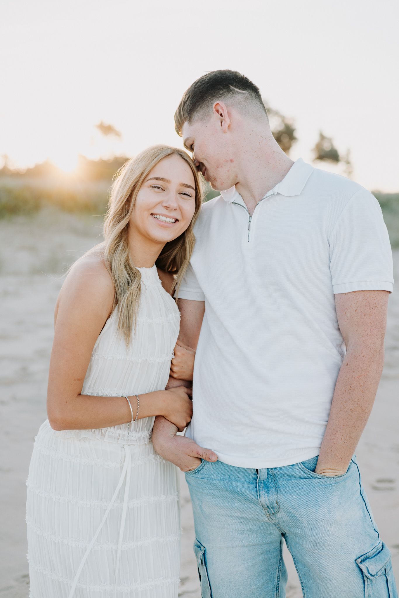 A smiling couple in white outfits embrace on a sandy beach at golden hour sunset.