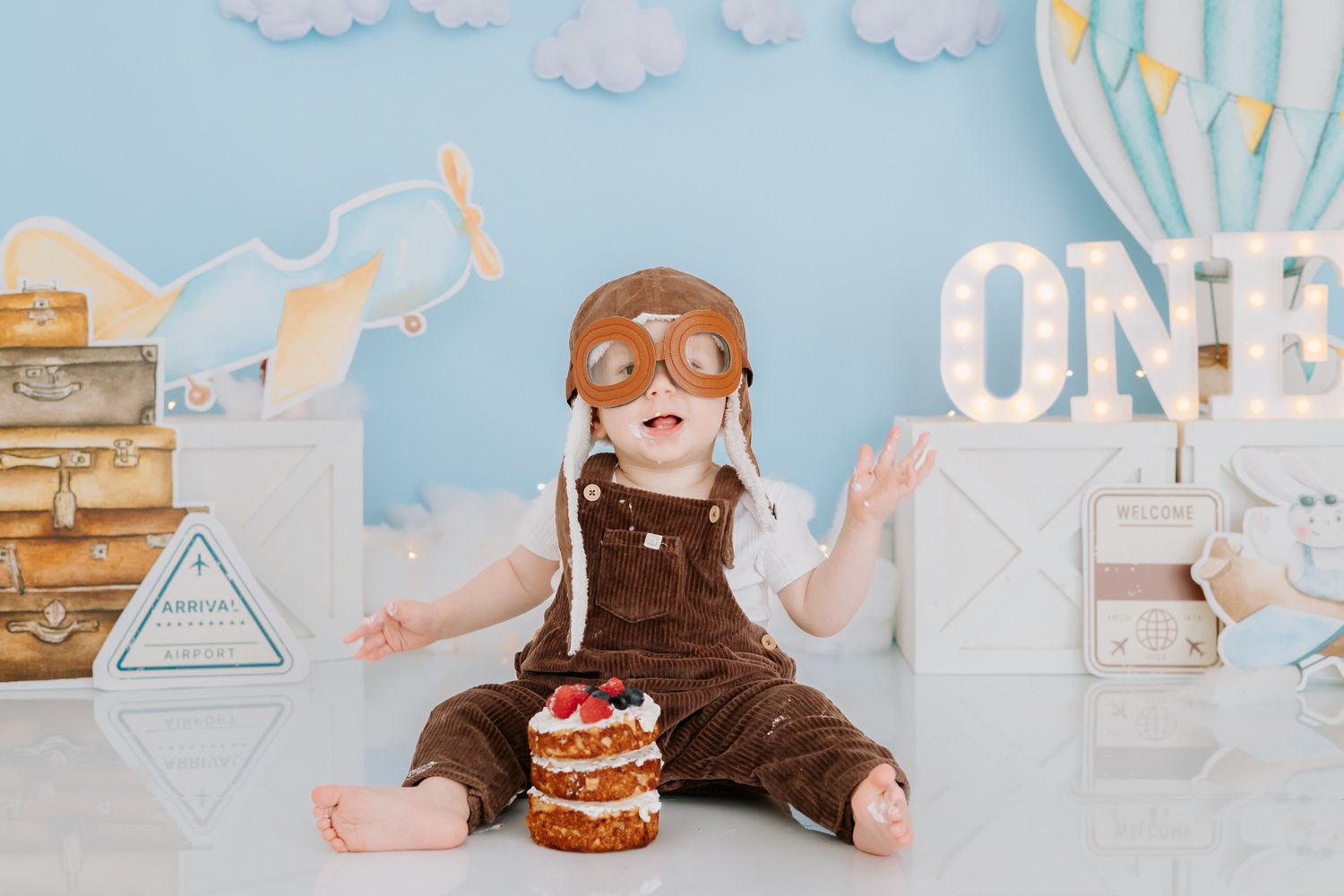 Baby in aviator hat sits with smash cake celebrating first birthday, surrounded by travel-themed decorations and ONE sign.