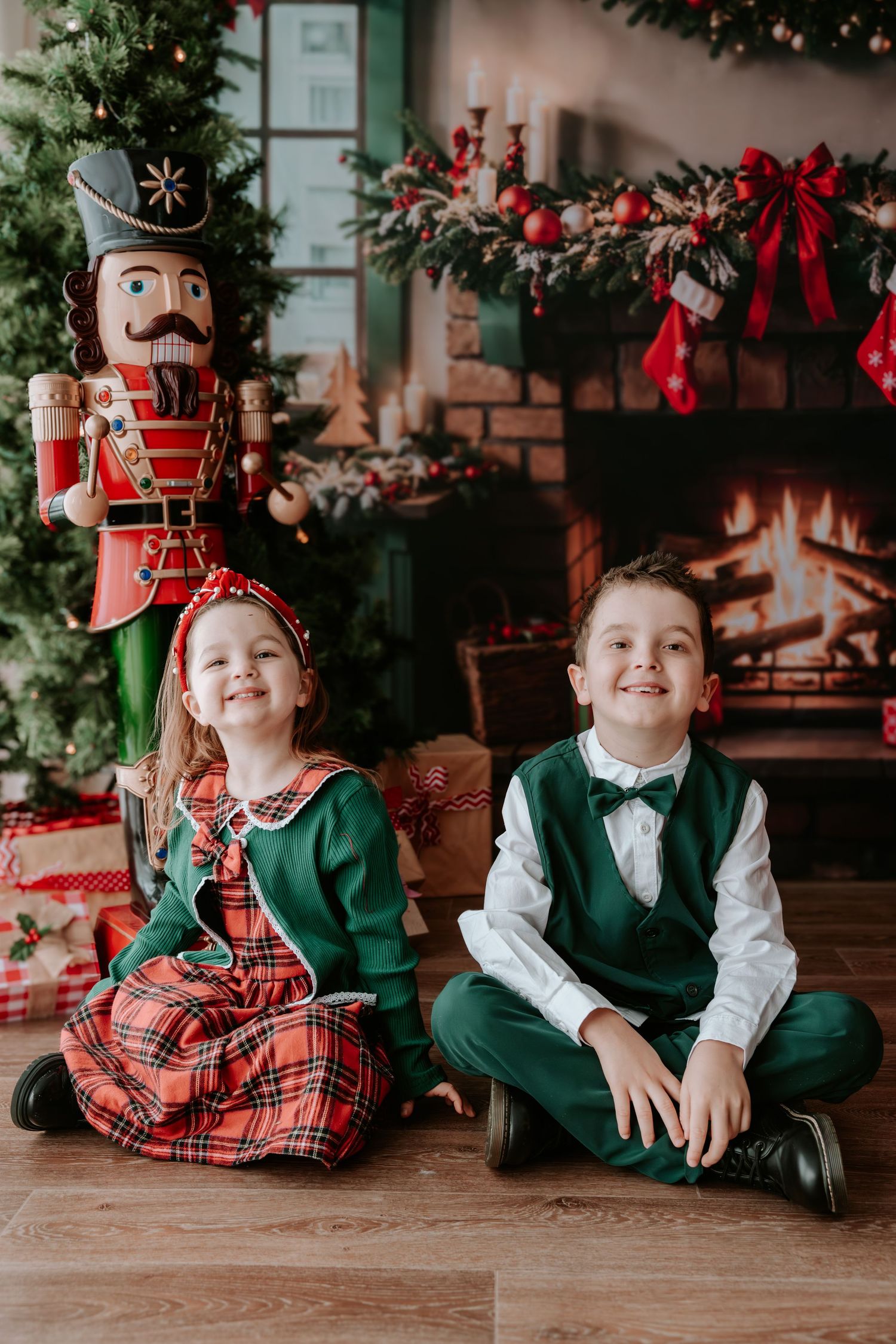 Two children in festive green outfits sit smiling by a decorated Christmas tree and fireplace with a nutcracker.