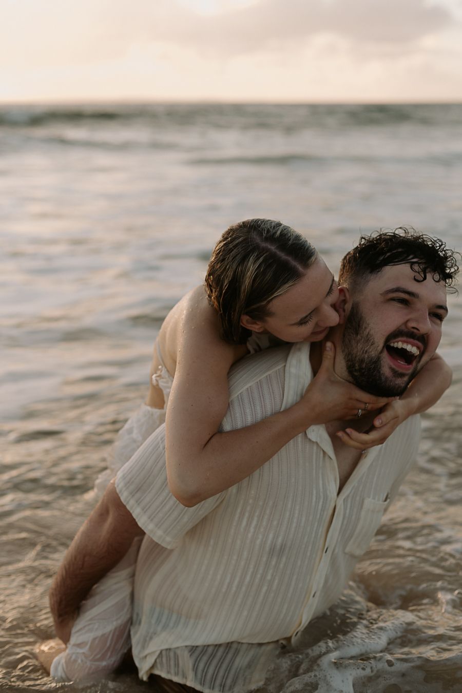 Couple laughing and playing in ocean waves at sunset, man giving woman a piggyback ride on the beach.