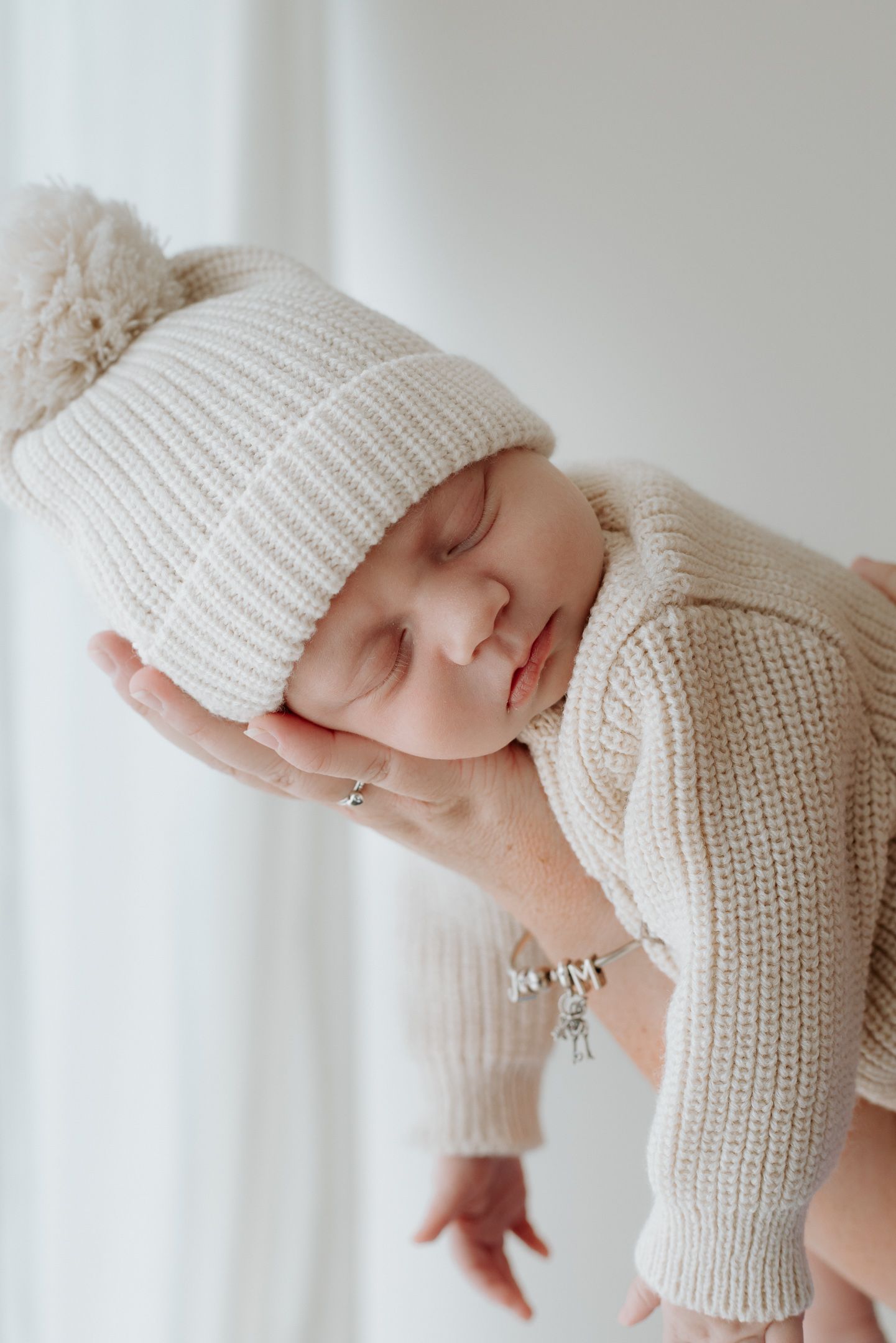 A sleeping newborn wearing a cozy white knit outfit and hat lies peacefully in a bright, minimalist setting.