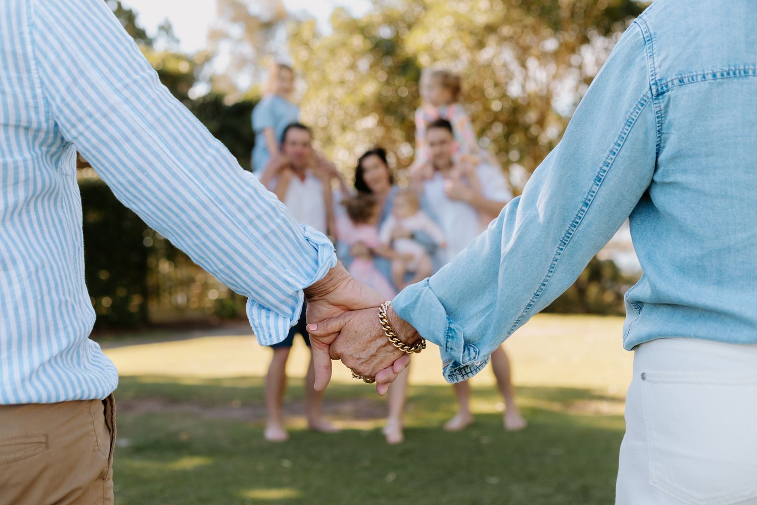 Couple holding hands in matching denim shirts with family group celebrating in sunny park background.