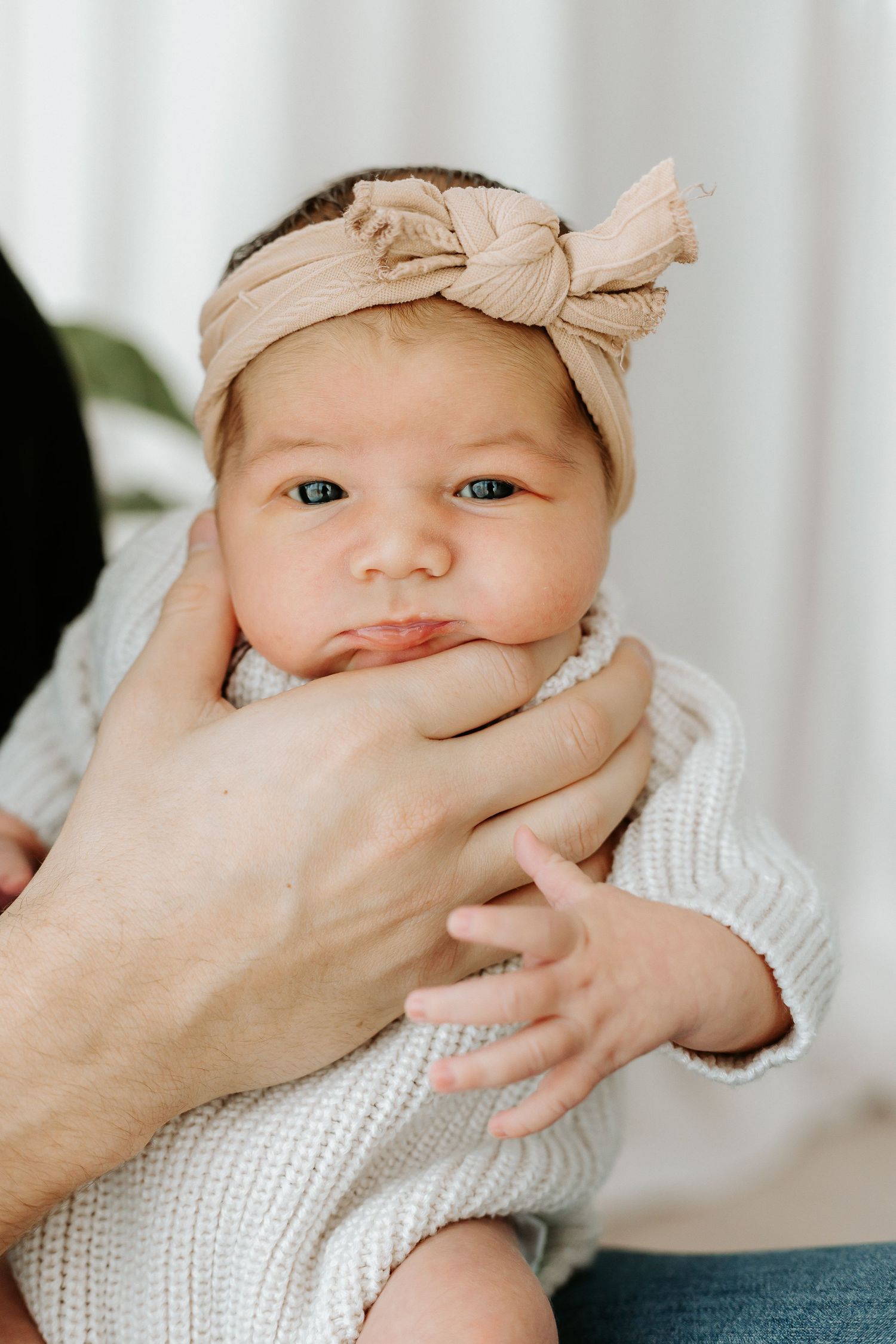 Newborn baby girl with blue eyes wearing a beige bow headband, cradled gently in adult hands.