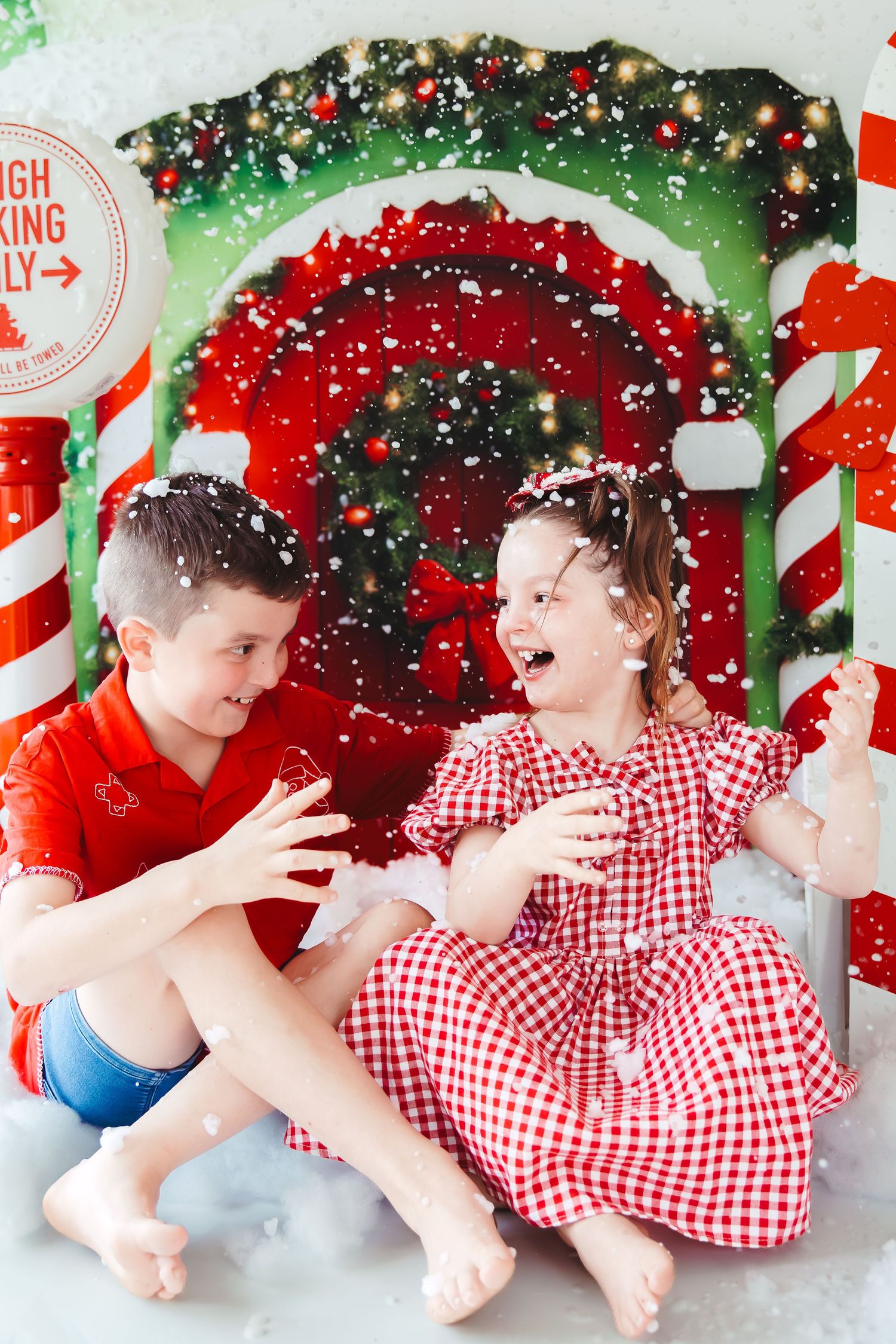 Two children laugh and play in artificial snow beside a festive Christmas candy cane backdrop.