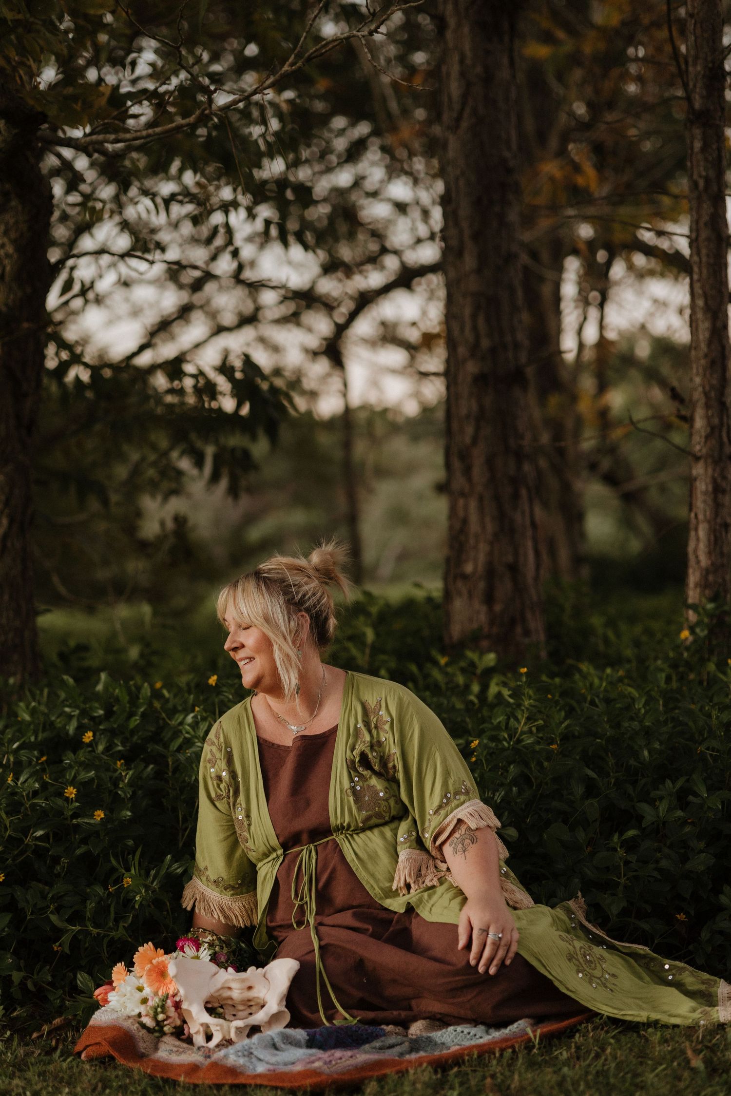 Woman in green kimono sitting peacefully outdoors among trees, smiling softly with flowers beside her.