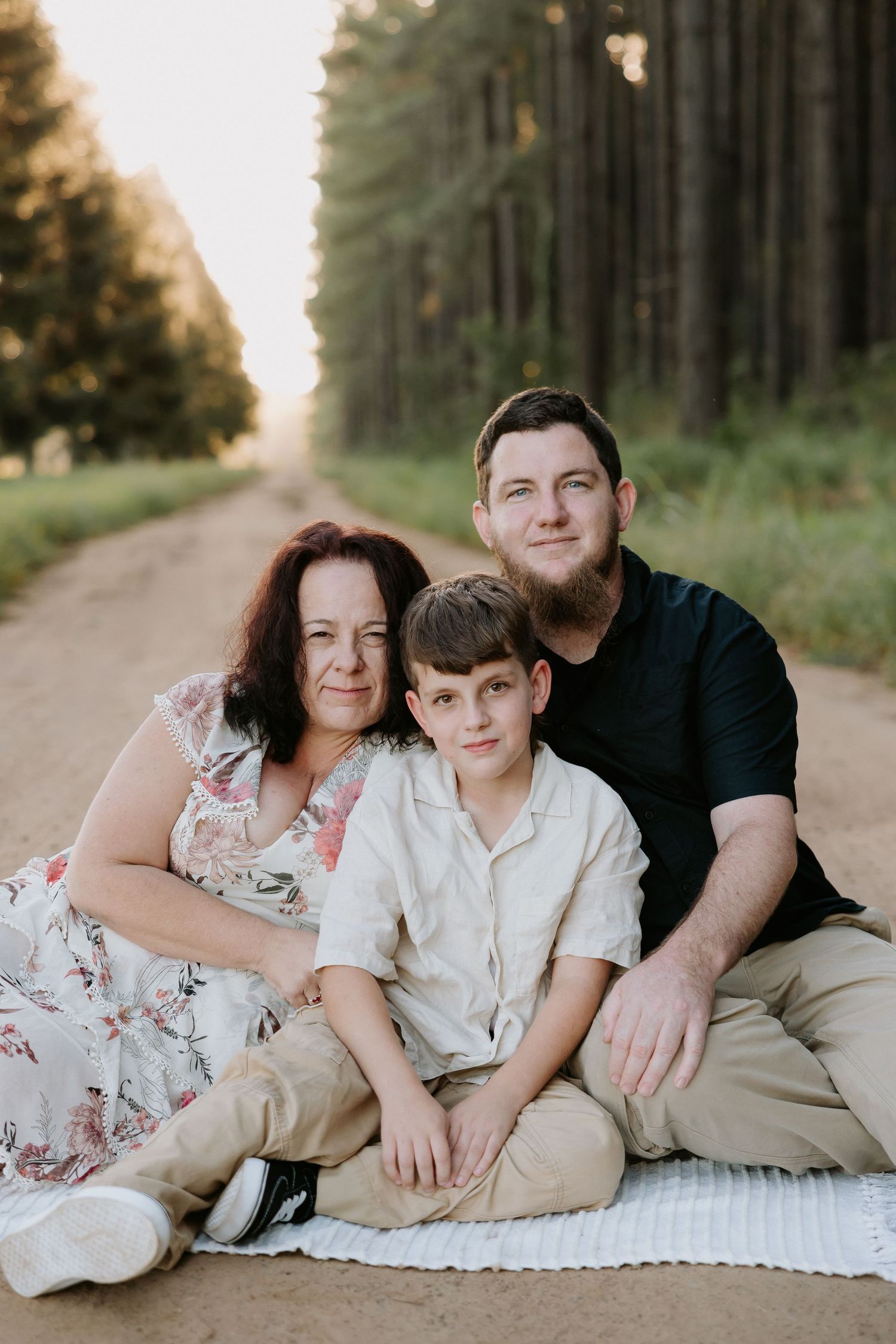 Family of three sitting on a blanket on a forest dirt road, smiling warmly in golden hour light.