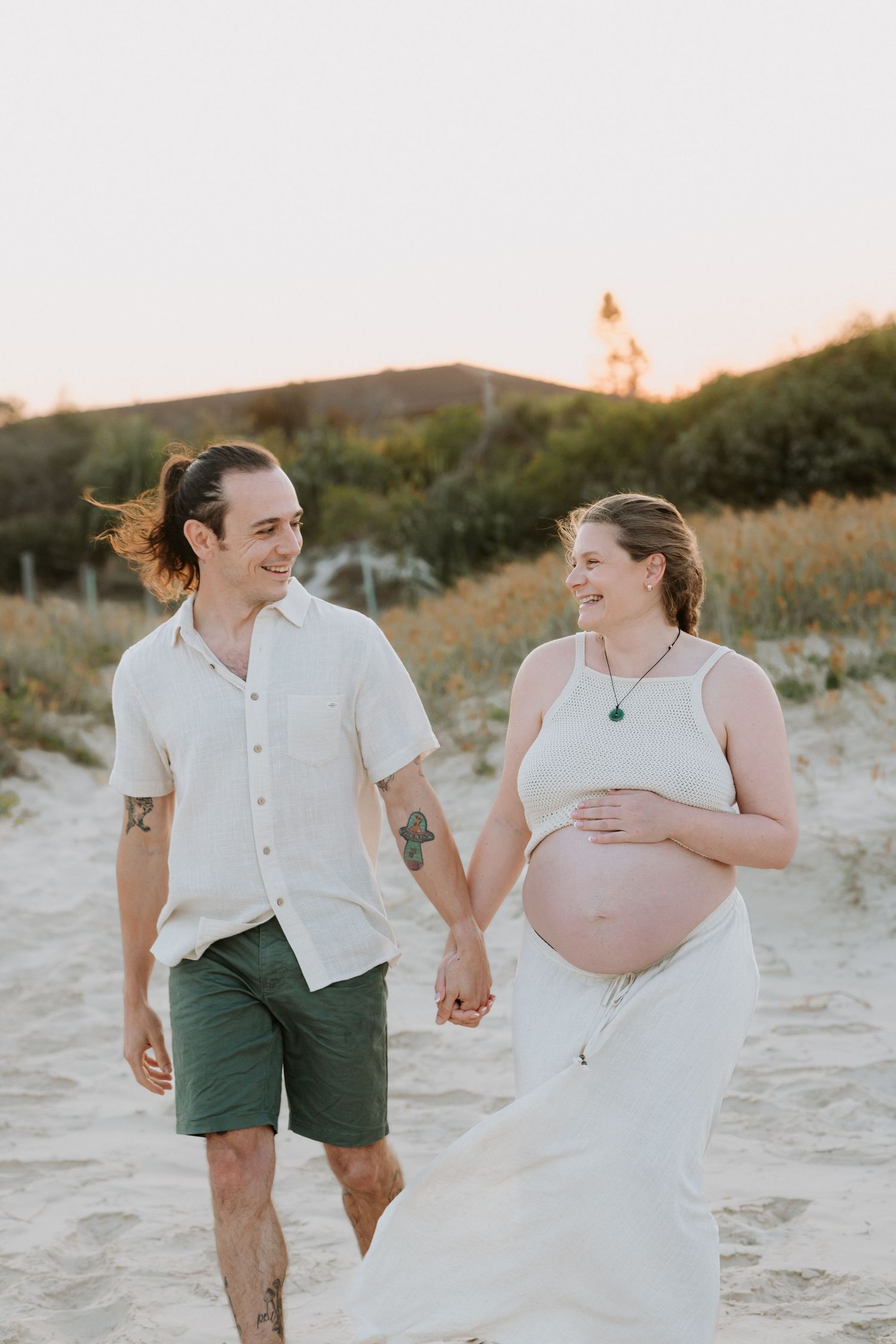 A couple walks hand in hand on a sandy beach at sunset, with a lighthouse visible in the background.
