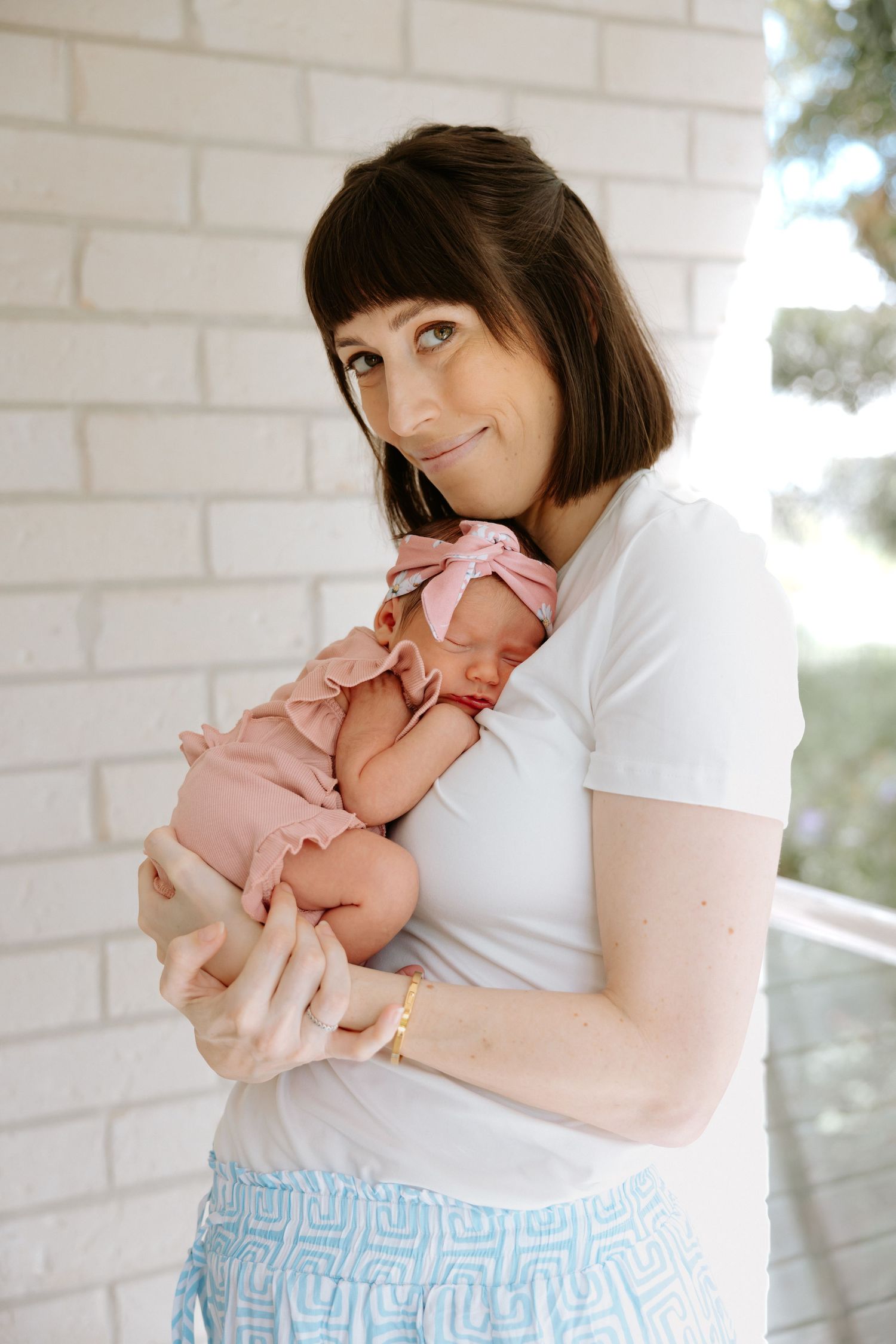 Parent in white shirt and blue patterned skirt holds newborn baby wearing pink outfit against white brick wall.