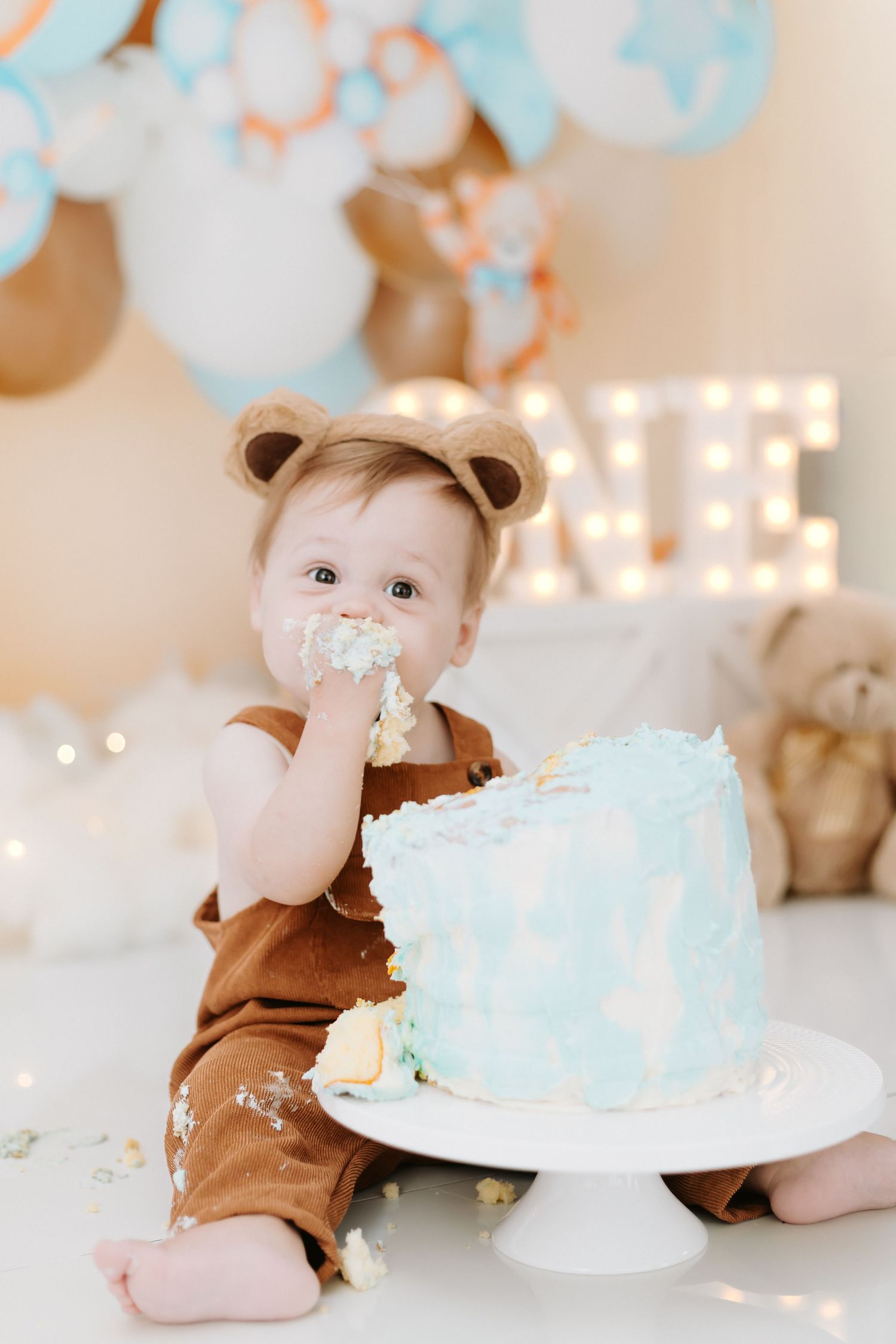 Baby in brown bear costume smashes face into light blue birthday cake during first birthday cake smash photoshoot.