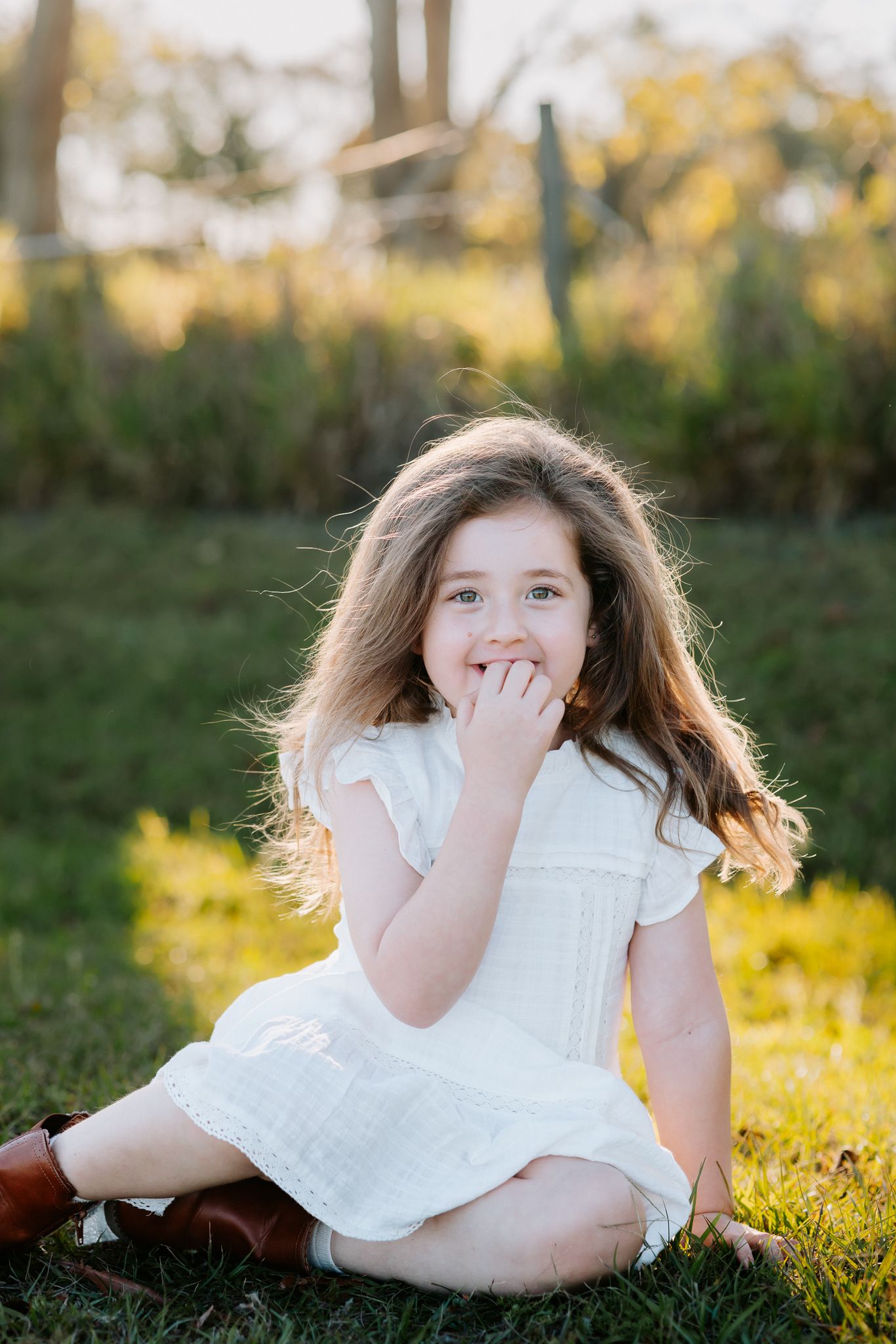 Young girl in white dress sitting on grass outdoors, smiling with hand over mouth in golden sunlight.