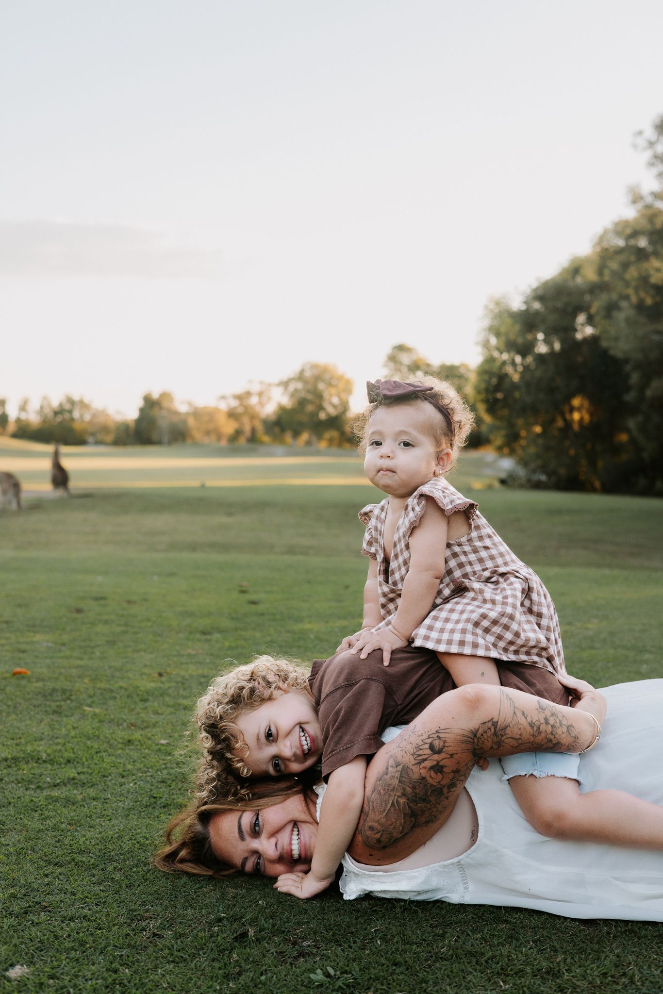 A mother and two children playing on green grass in a park during golden hour, laughing and stacking on top of each other.