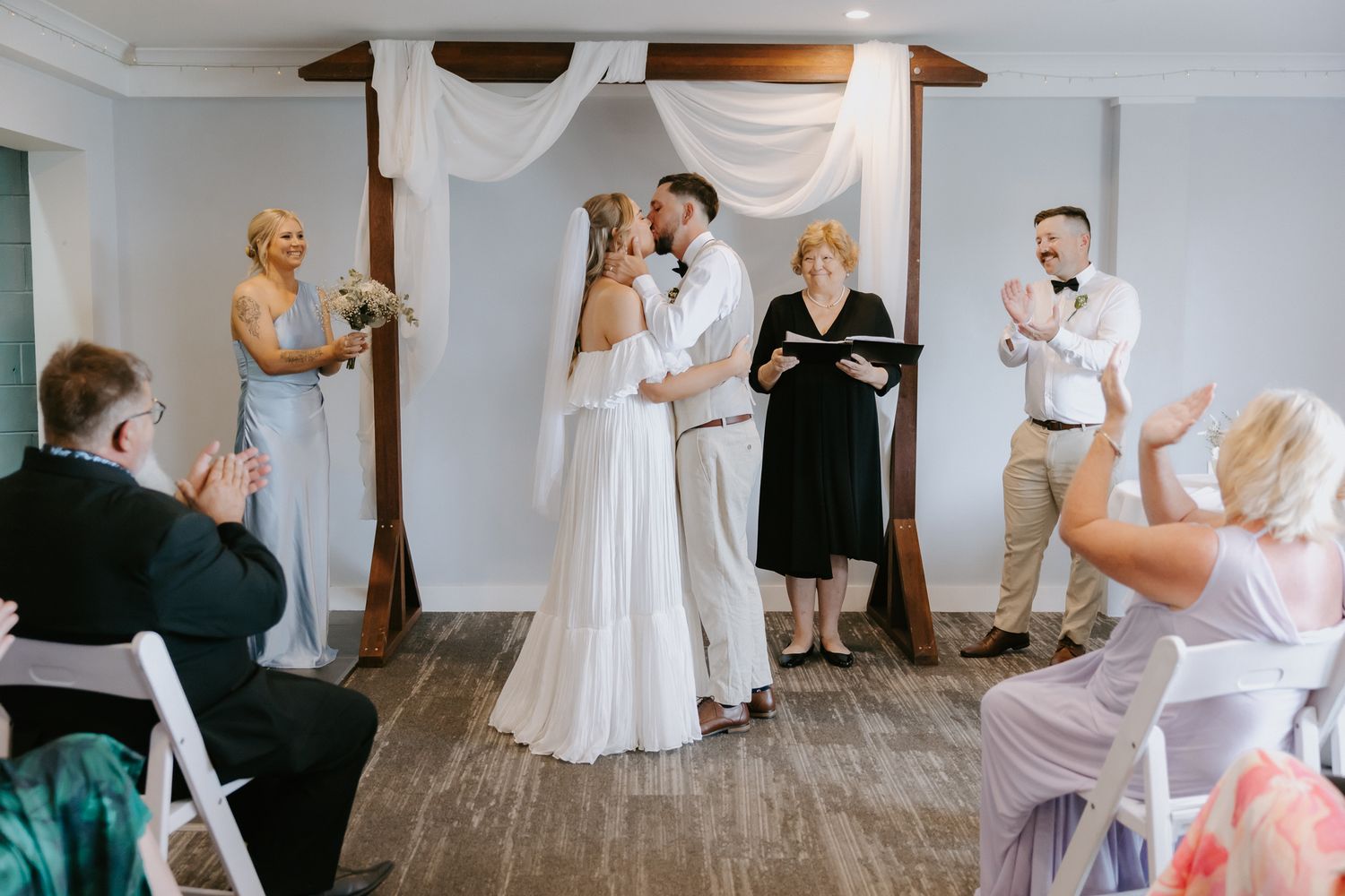 Bride and groom share first kiss at altar surrounded by clapping guests during indoor wedding ceremony.