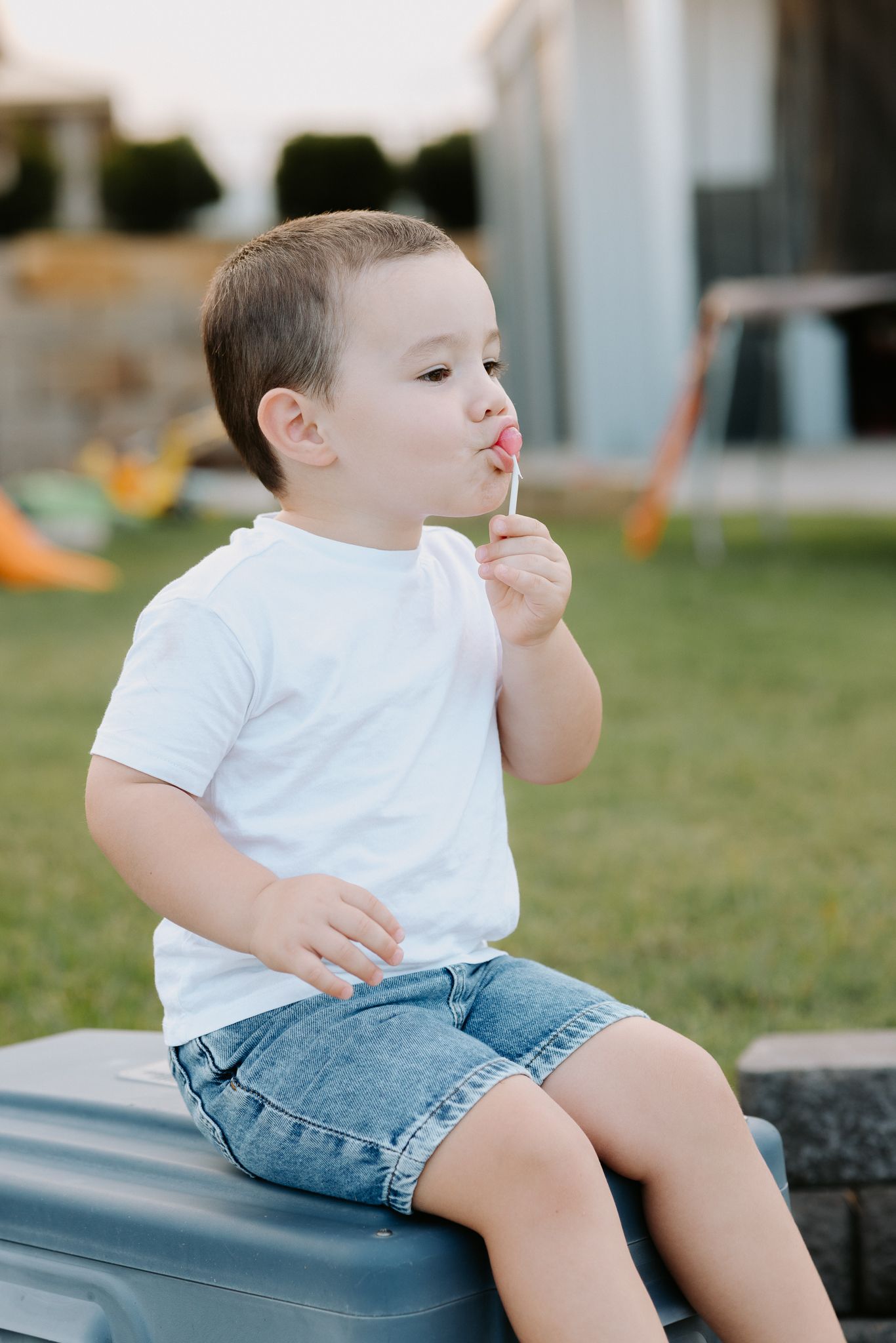 Cute toddler boy in white t-shirt and denim shorts eating a lollipop outdoors.