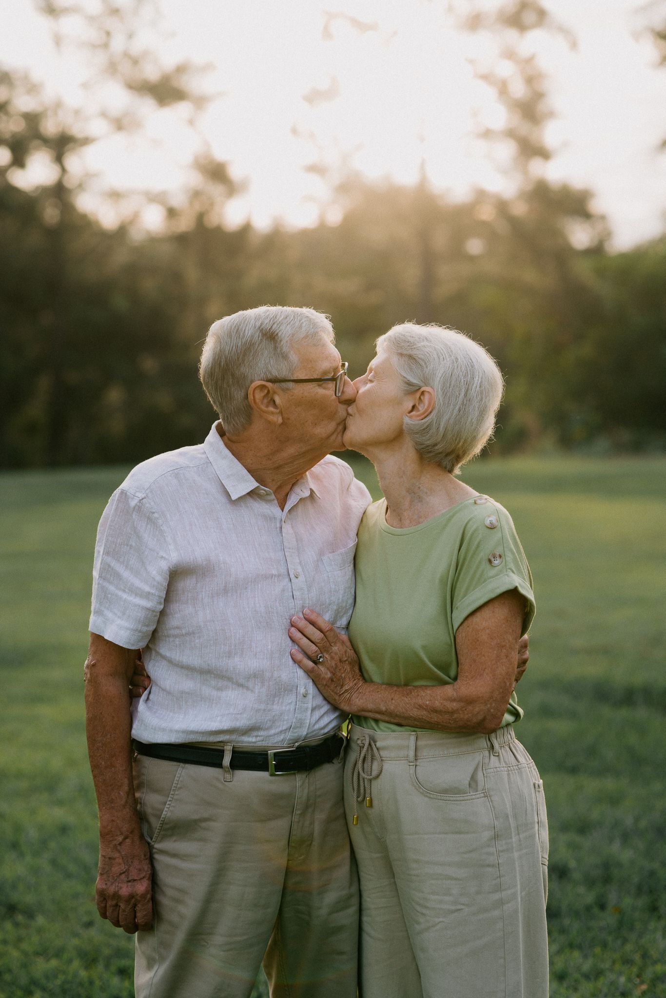 Elderly couple sharing a tender kiss outdoors on a green field at sunset, wearing casual light-colored clothing.