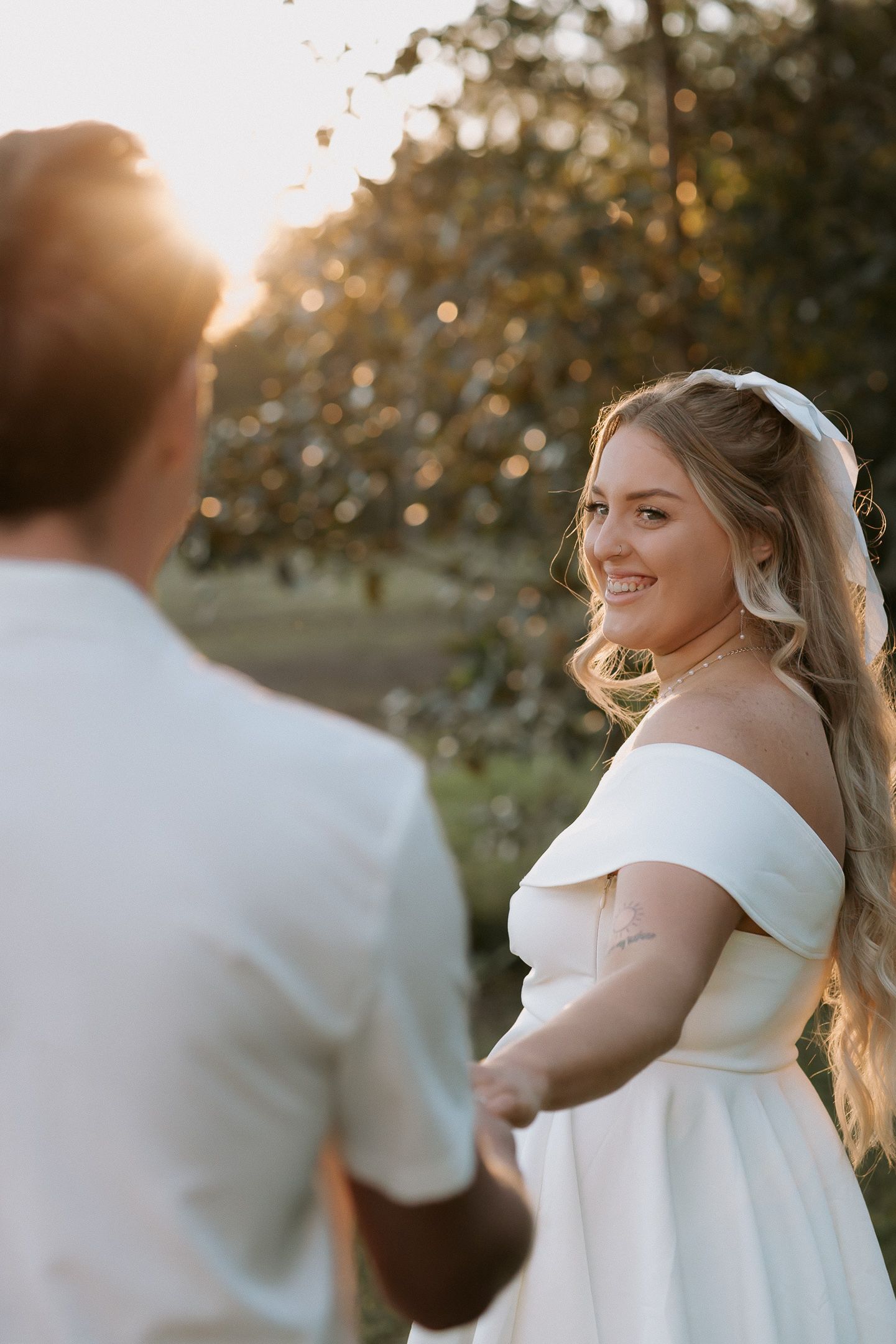 Bride in off-shoulder white gown holds hands with groom at golden hour sunset outdoors.