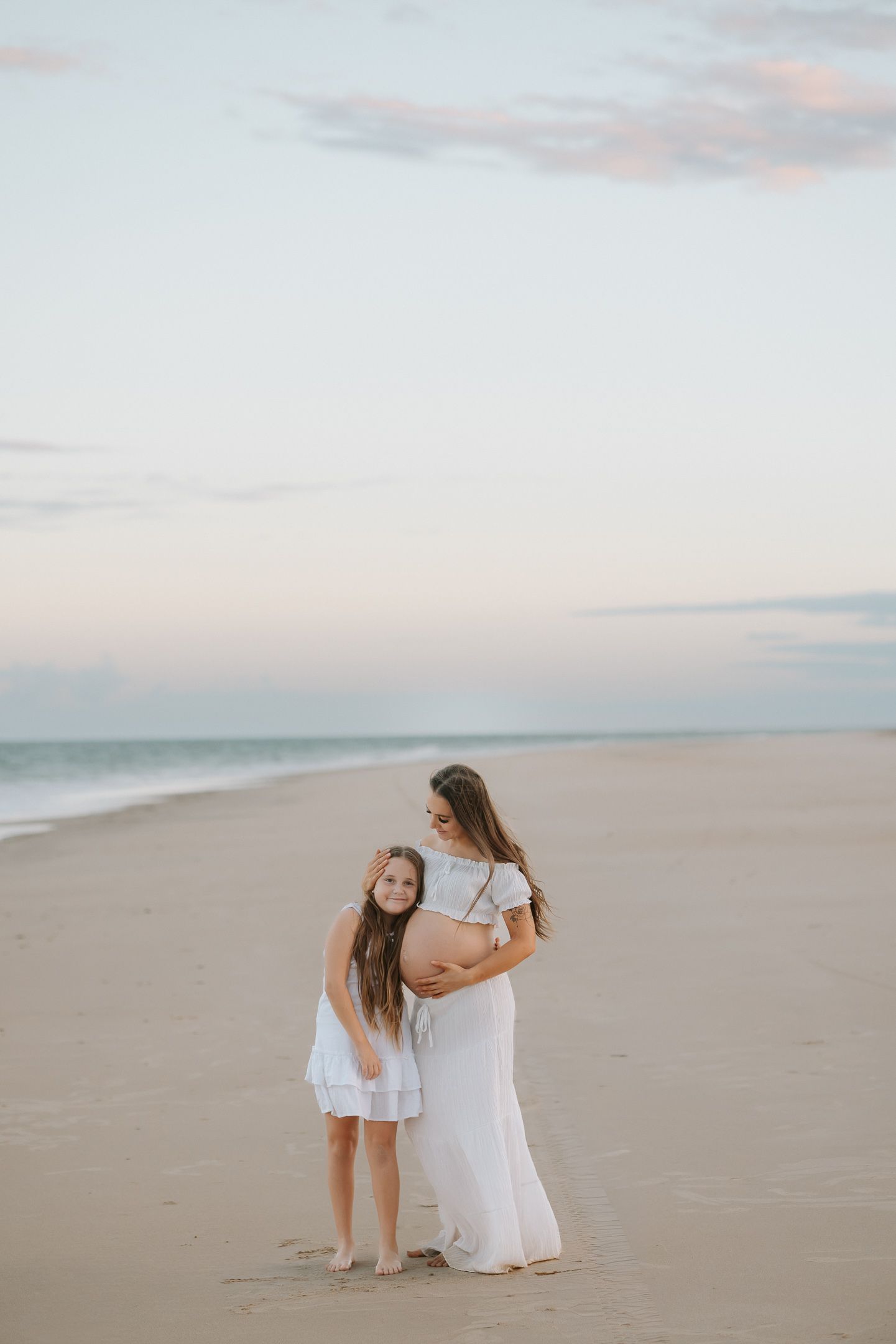 Two people in white dresses sit together on a sandy beach at sunset creating a dreamy coastal scene.