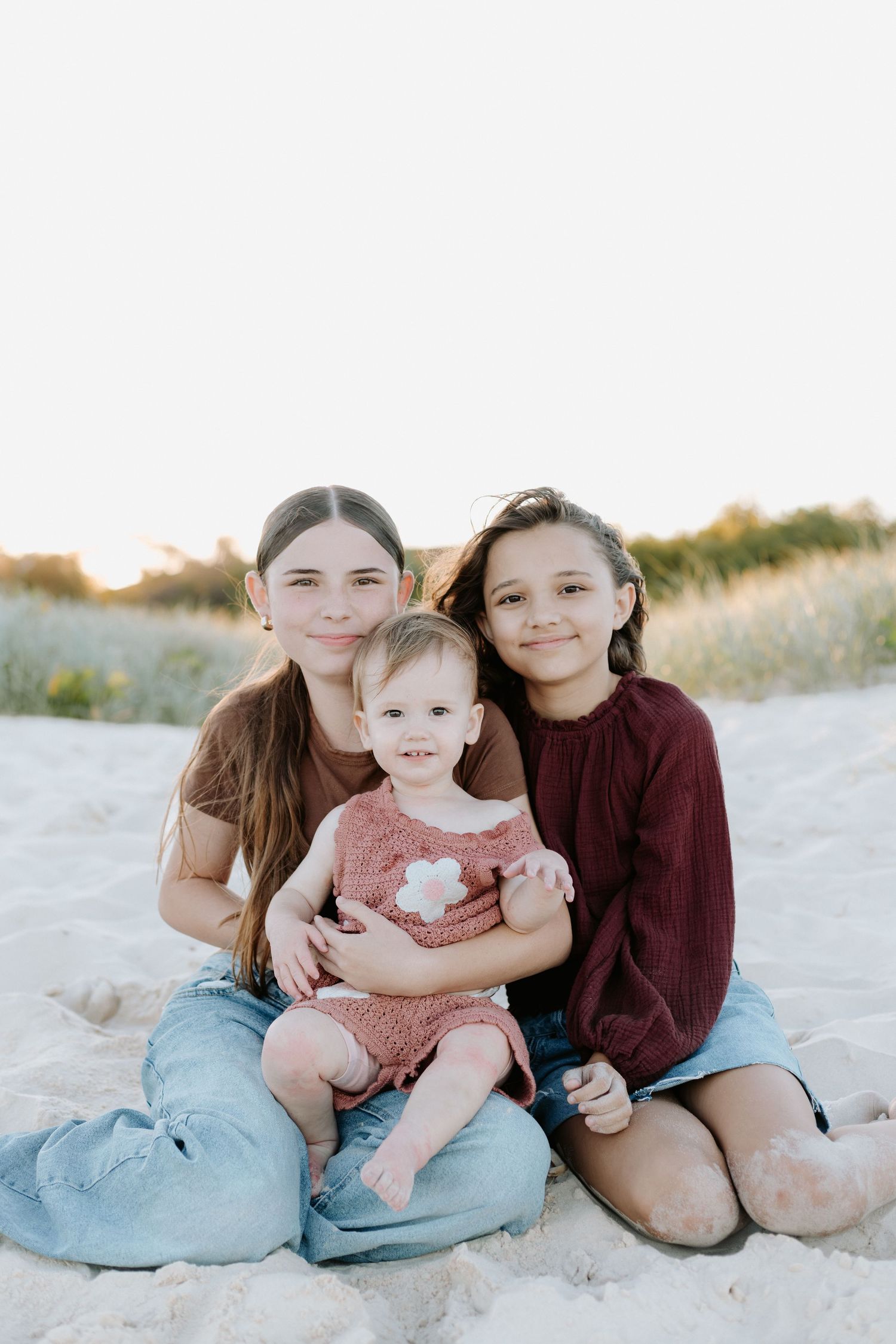 A family sits together on a sandy beach during sunset in matching burgundy and pink outfits.