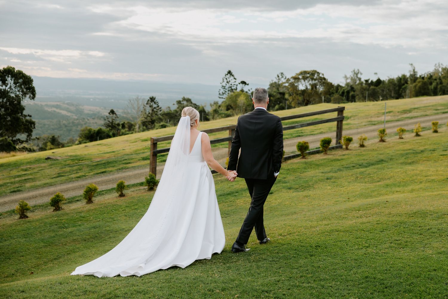 Bride and groom walk hand in hand across lush green hillside with scenic countryside views and wooden fence backdrop.