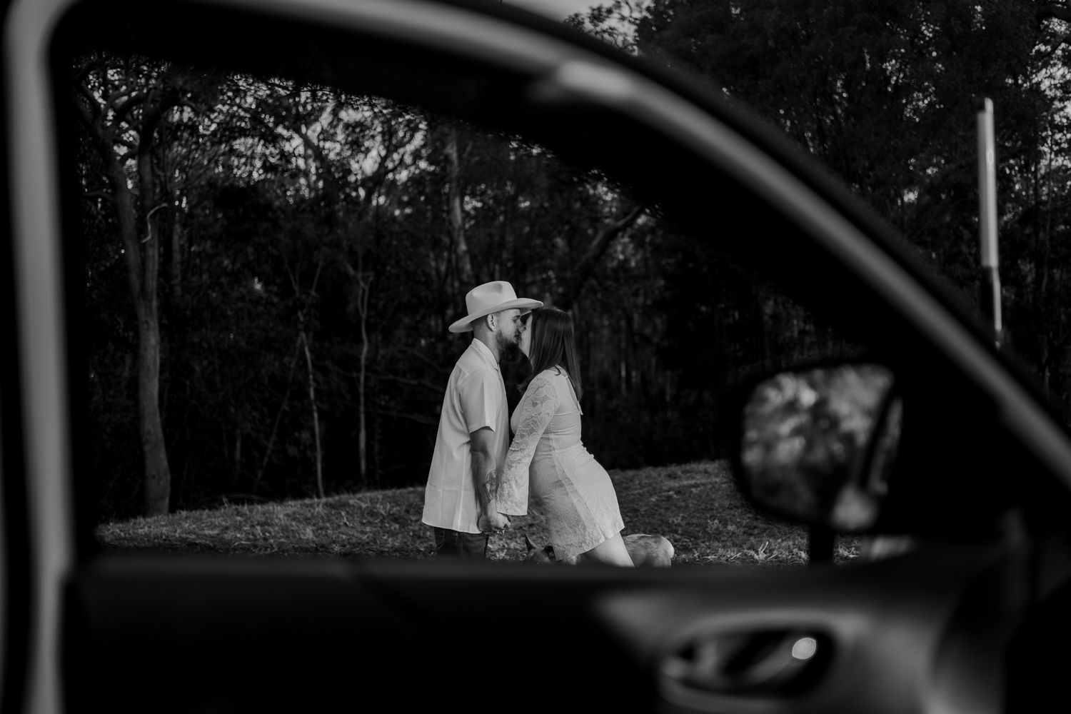 Black and white photo of a couple kissing outdoors, framed through a car window, with trees in the background.