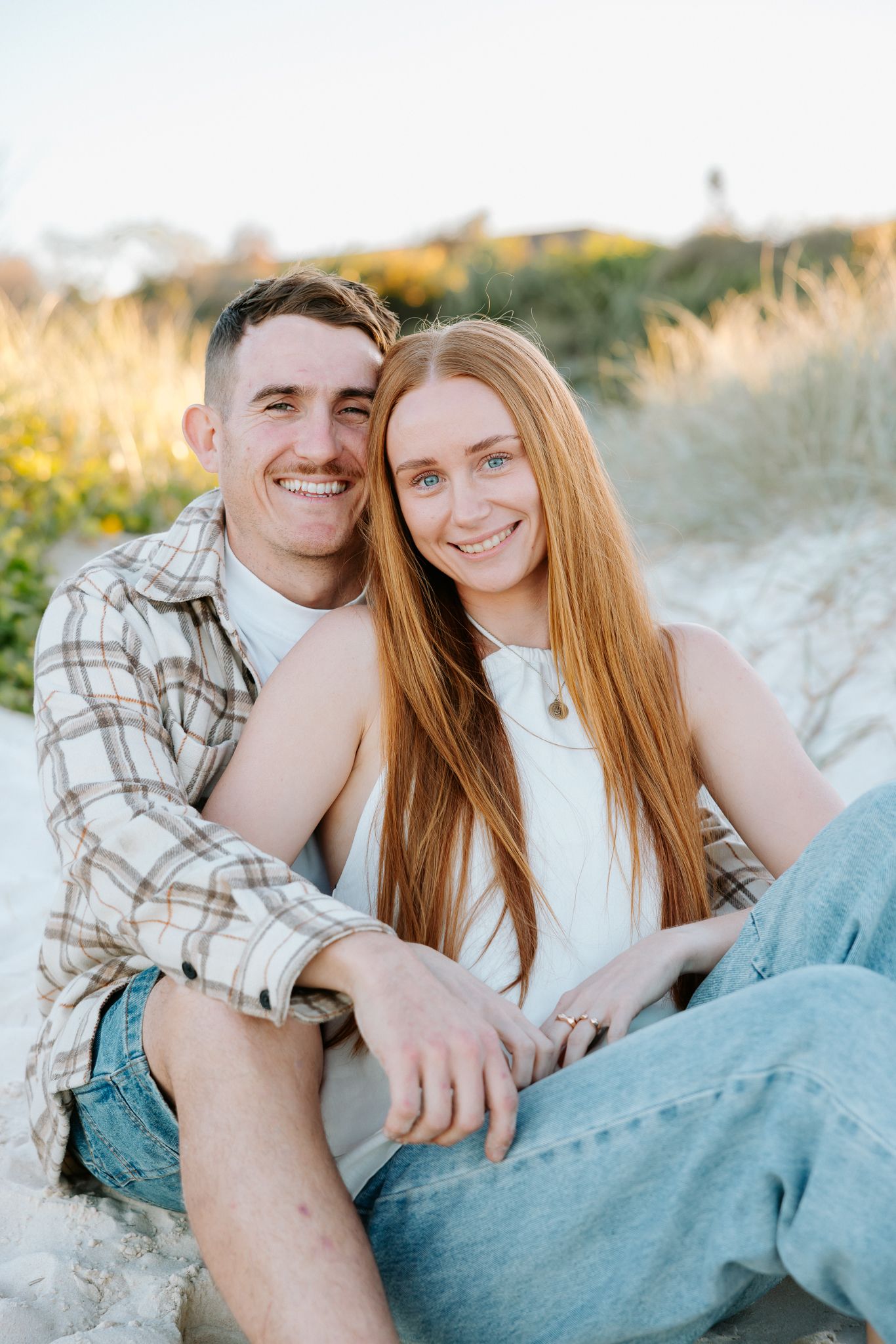 Happy young couple sitting on sandy beach dunes, woman with long red hair in white top, man in plaid shirt smiling.