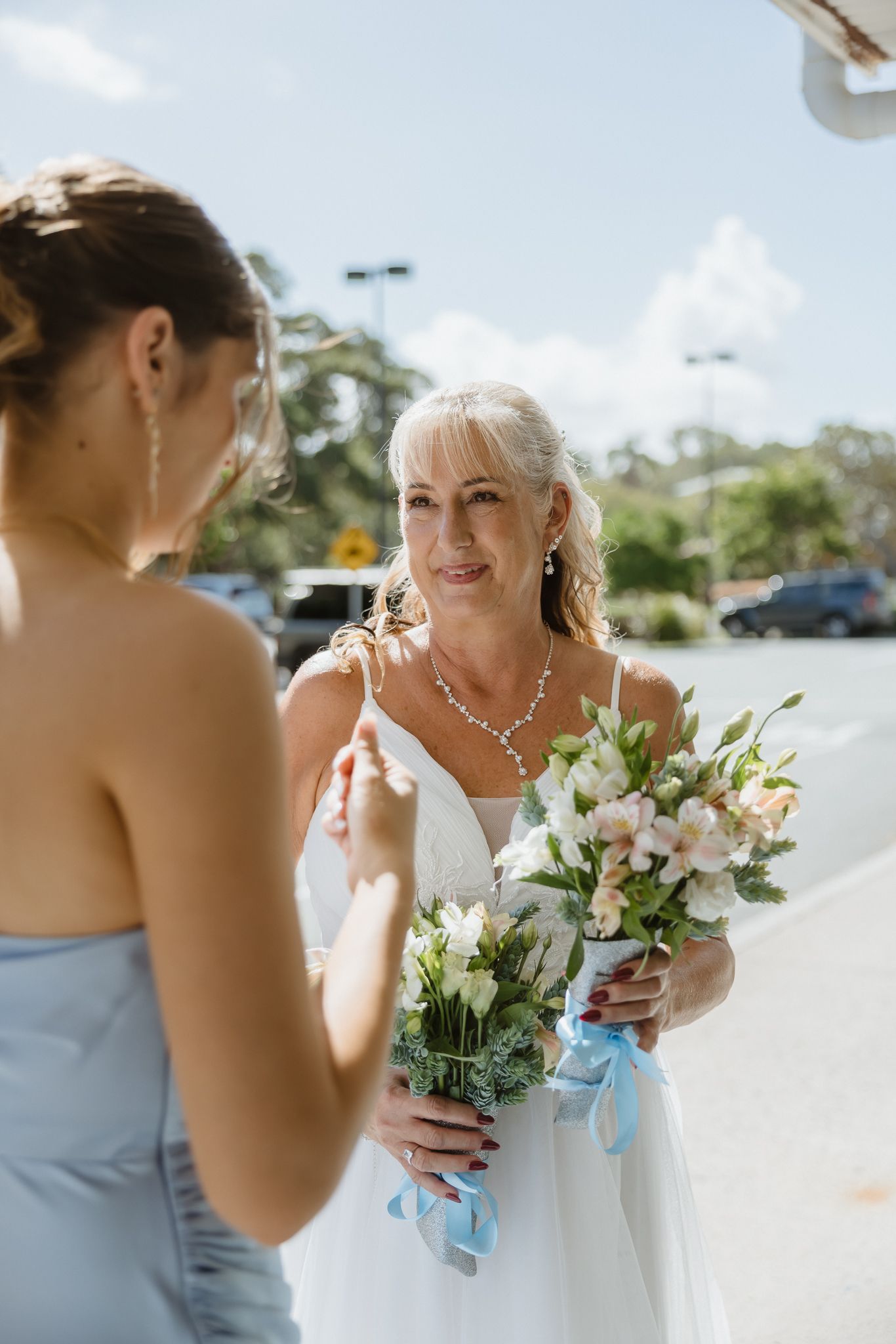Elegant bride in white gown holding bouquet of white and pink flowers talks with bridesmaid in blue dress outdoors.