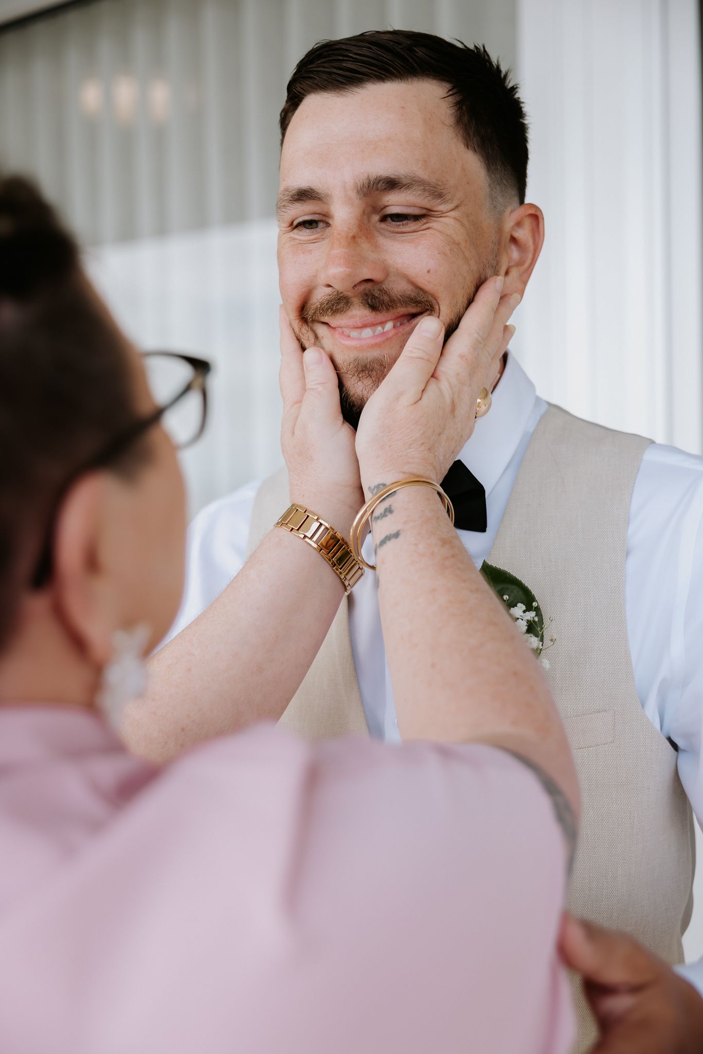 Smiling groom in beige vest receives tender face touch from woman during wedding day preparations.