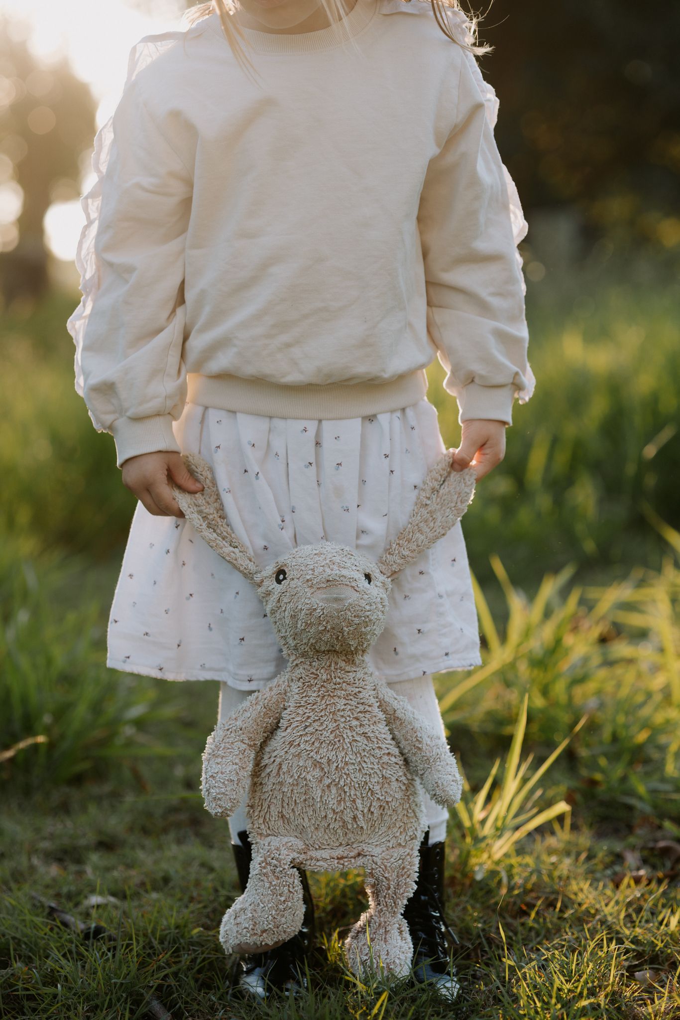 Young girl in white outfit holding a stuffed bunny toy while standing in a sunlit green field.