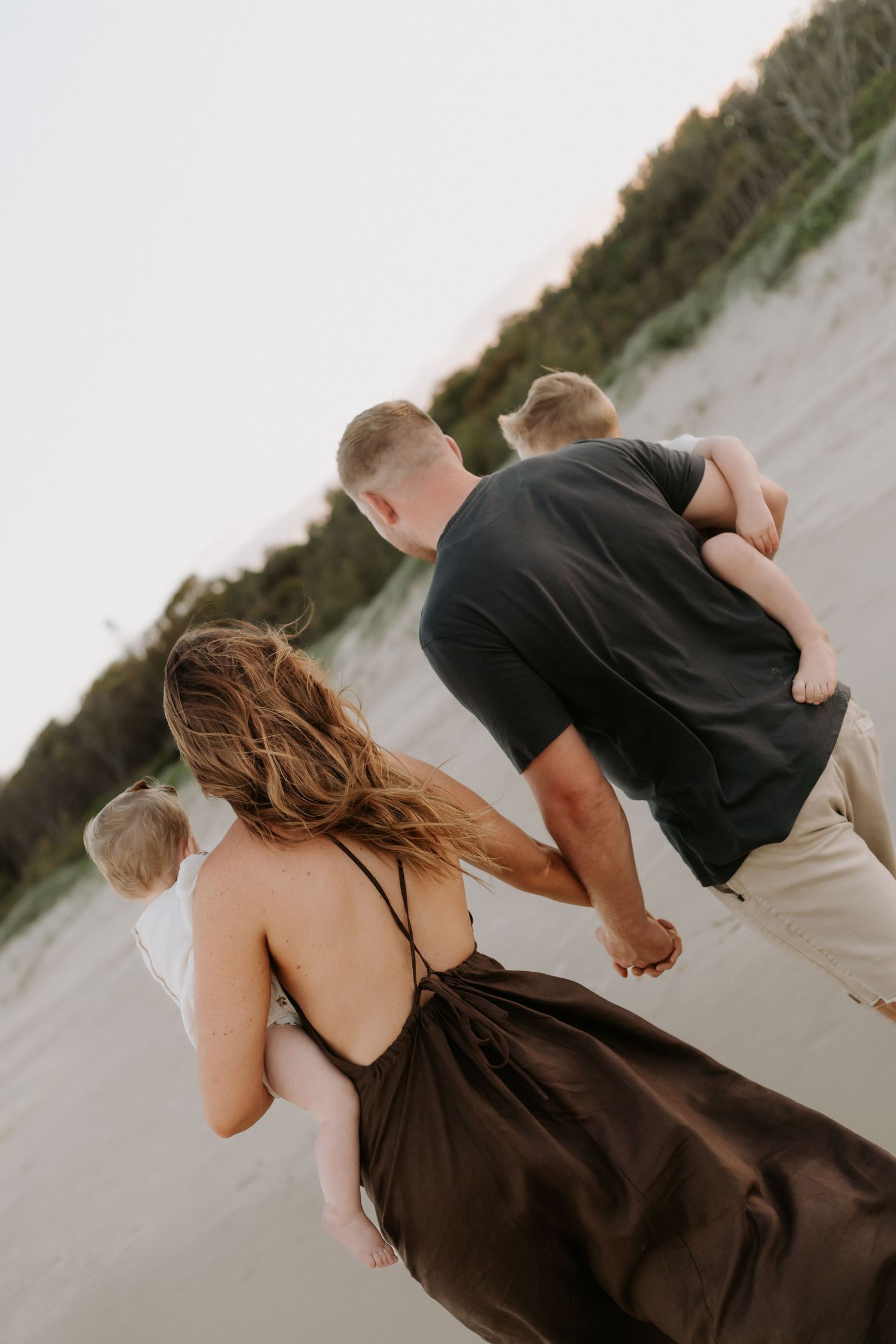 Family walking along a sandy beach, parents carrying young children, woman in brown dress holding hands with man in black shirt.