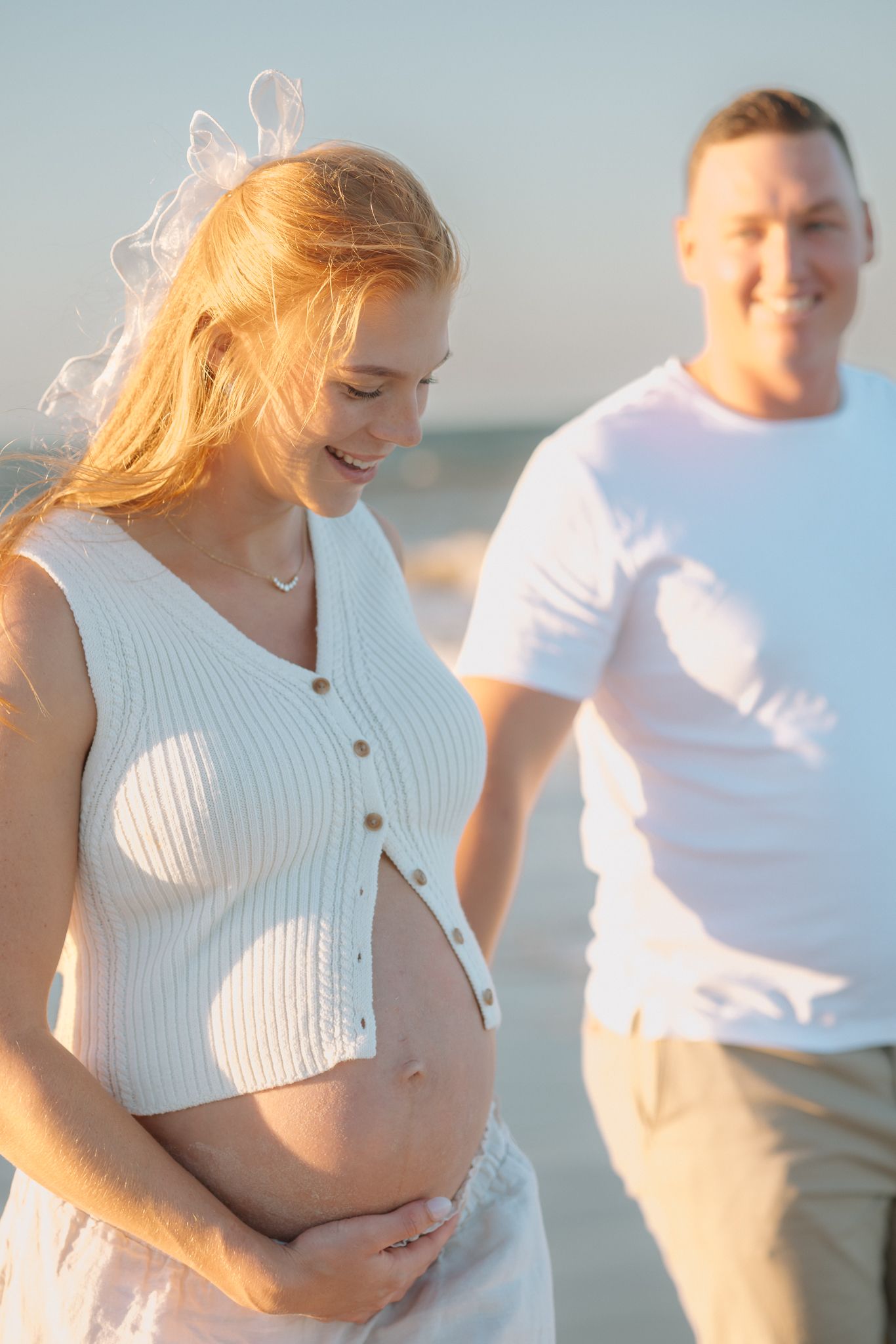 Pregnant woman in white top holds her baby bump while smiling, standing with her partner on a sunny beach.